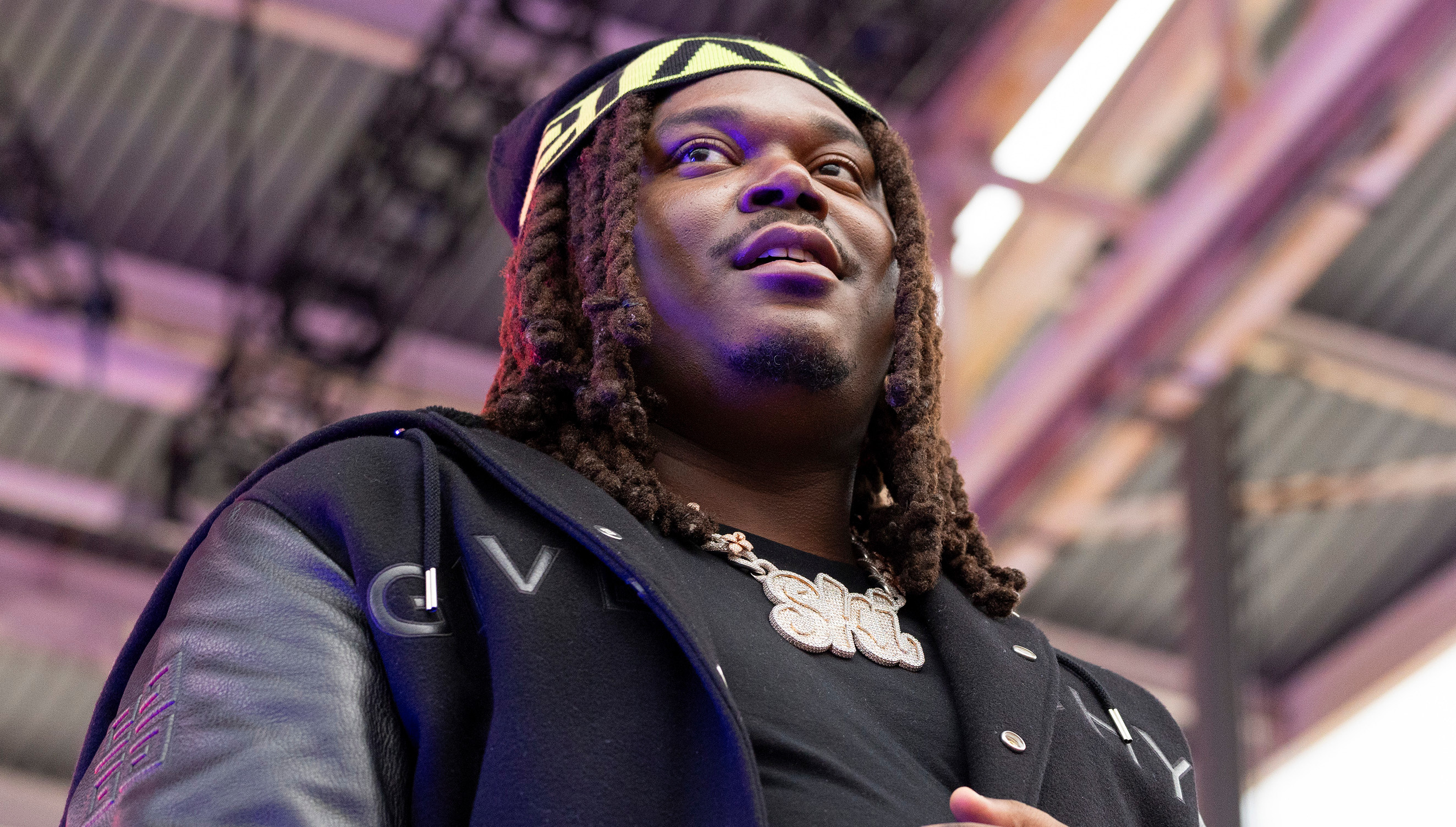 Lucki with long hair and a patterned headband, wearing a black jacket, stands under a metal structure, looking upward.
