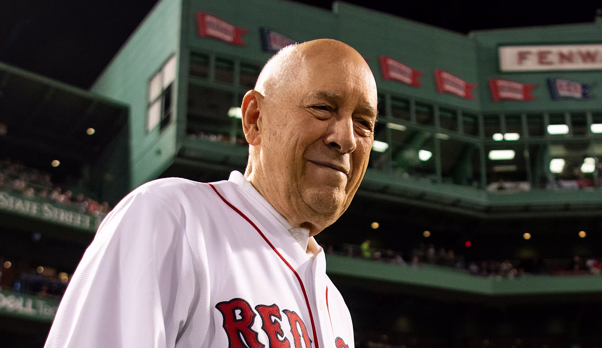 Bill Rasmussen in a Red Sox jersey stands at Fenway Park at night, with the stadium and crowd visible in the background.