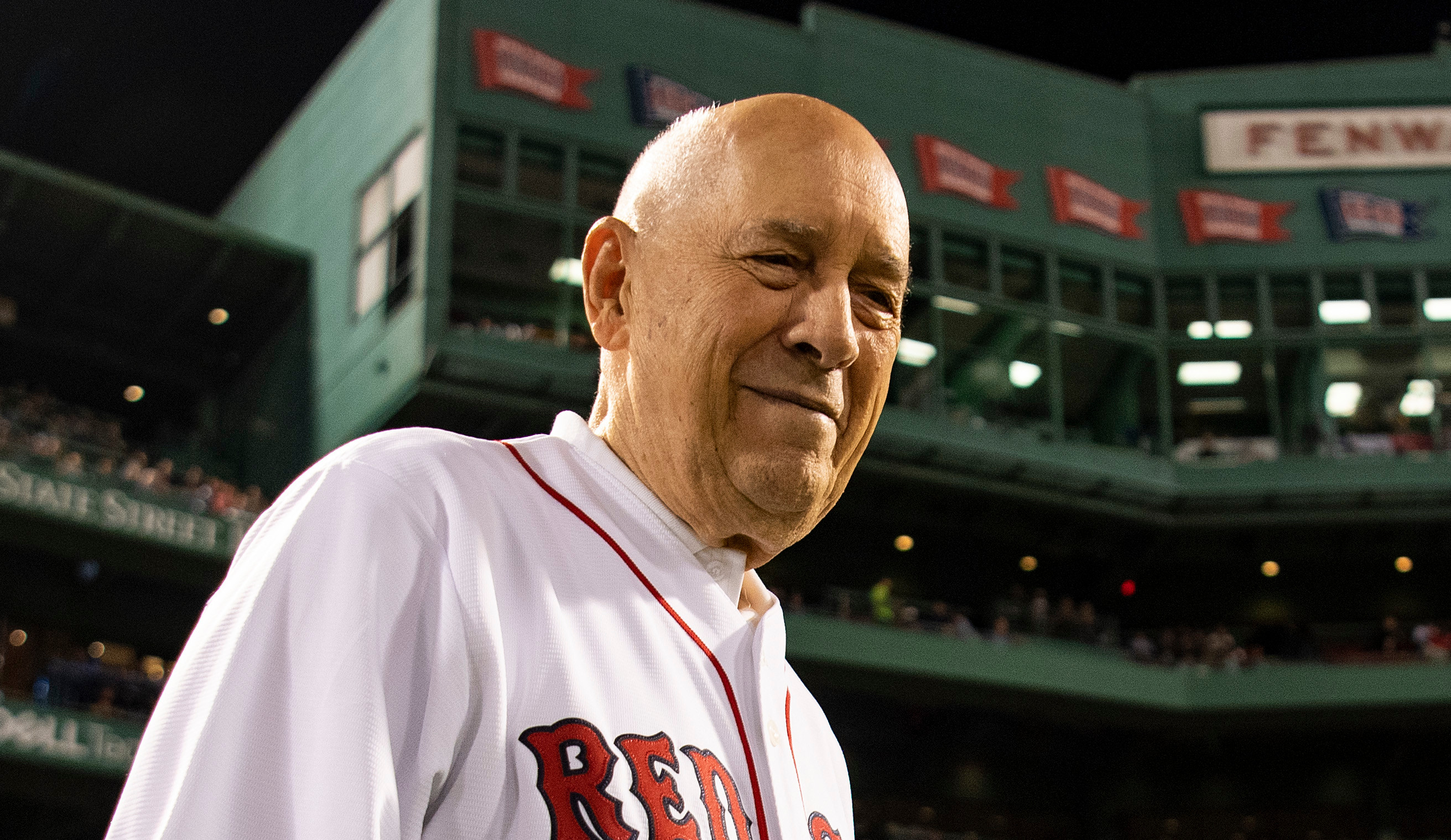 Bill Rasmussen in a Red Sox jersey stands at Fenway Park at night, with the stadium and crowd visible in the background.