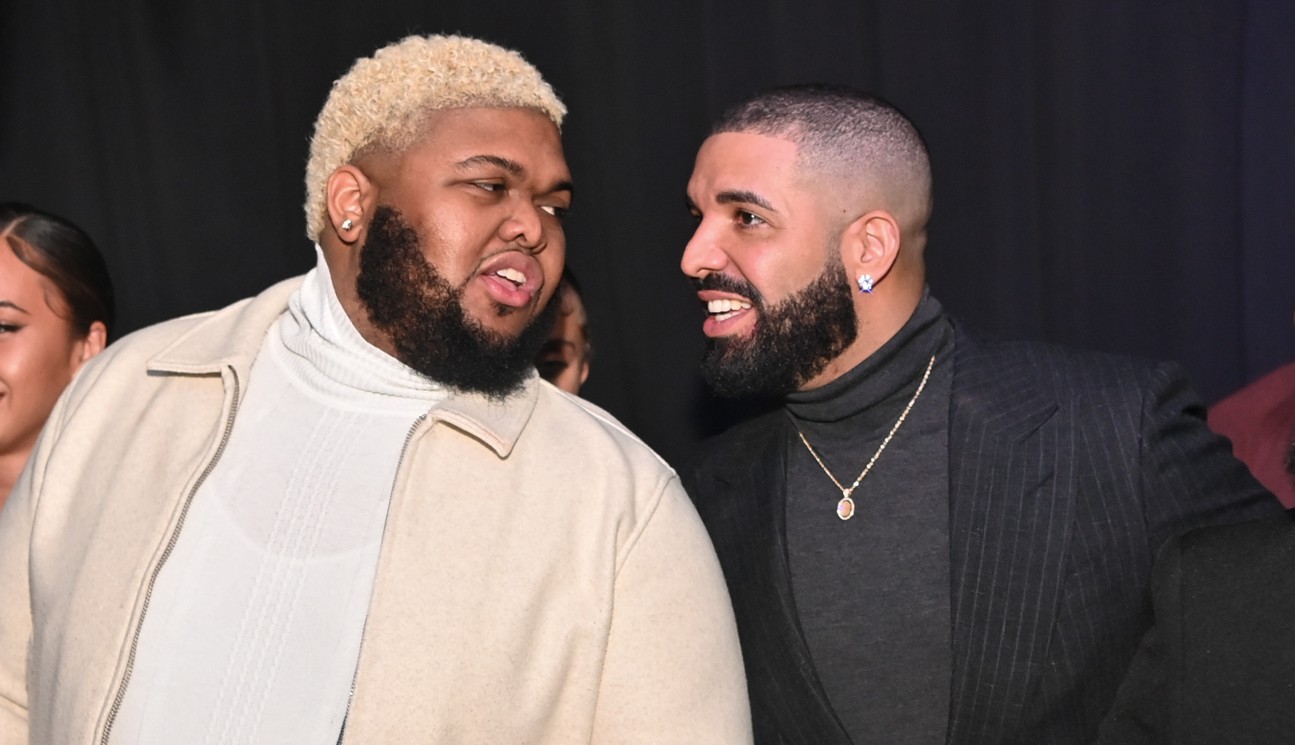 Drake, wearing a black turtleneck and necklace, converses with Druski in a white jacket at a social event.