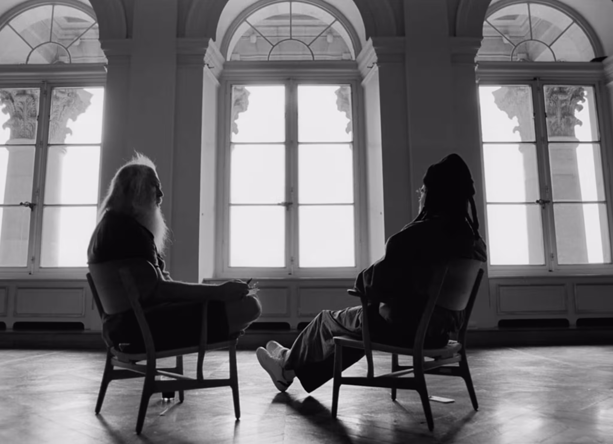 Jay-Z and Rick Rubin sit facing each other in chairs, silhouetted against large windows in a grand room.