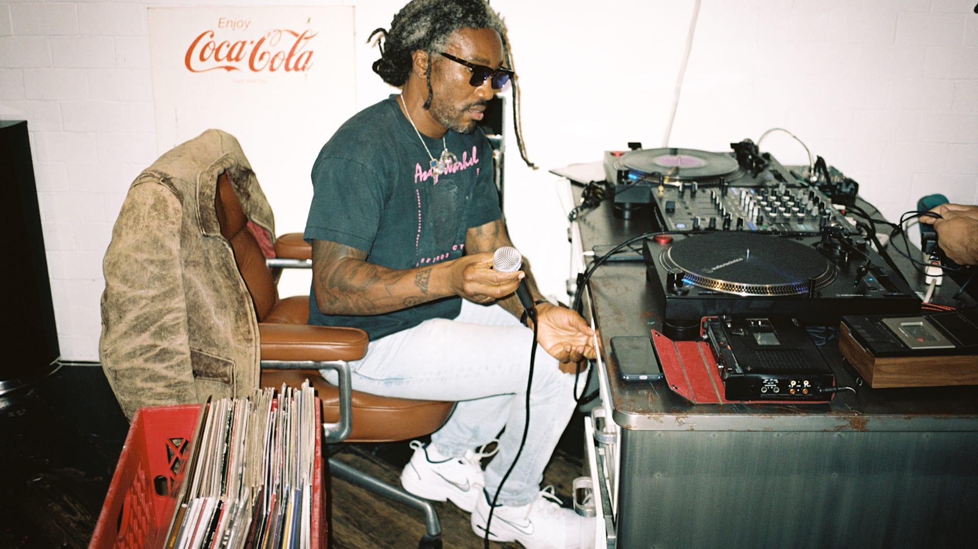 A person with sunglasses holding a microphone sits by DJ equipment, surrounded by vinyl records and a Coca-Cola sign in the background.