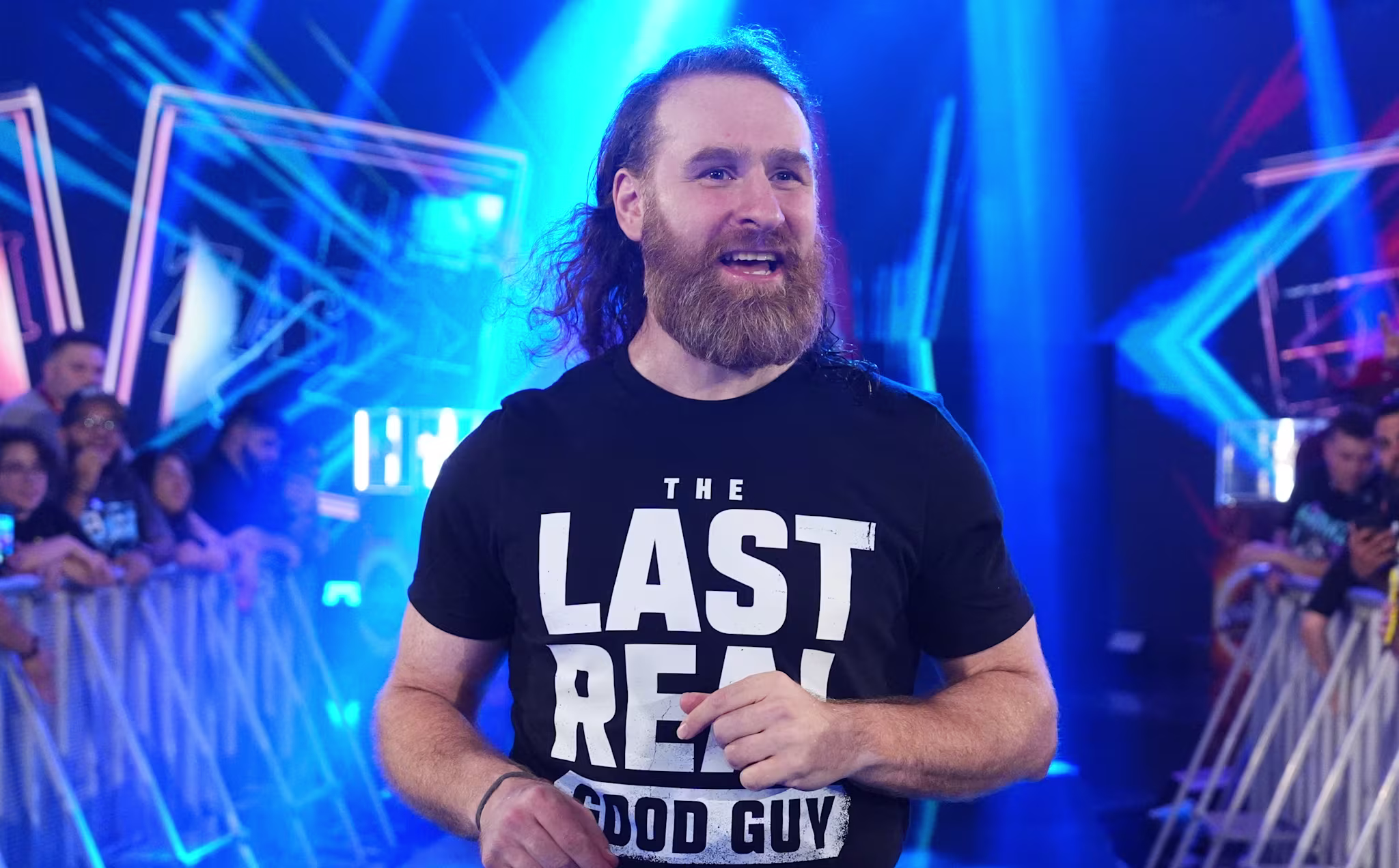 Sami Zayn with long hair and a beard walks down a lit aisle, wearing a shirt that says "The Last Real Good Guy," surrounded by cheering fans.