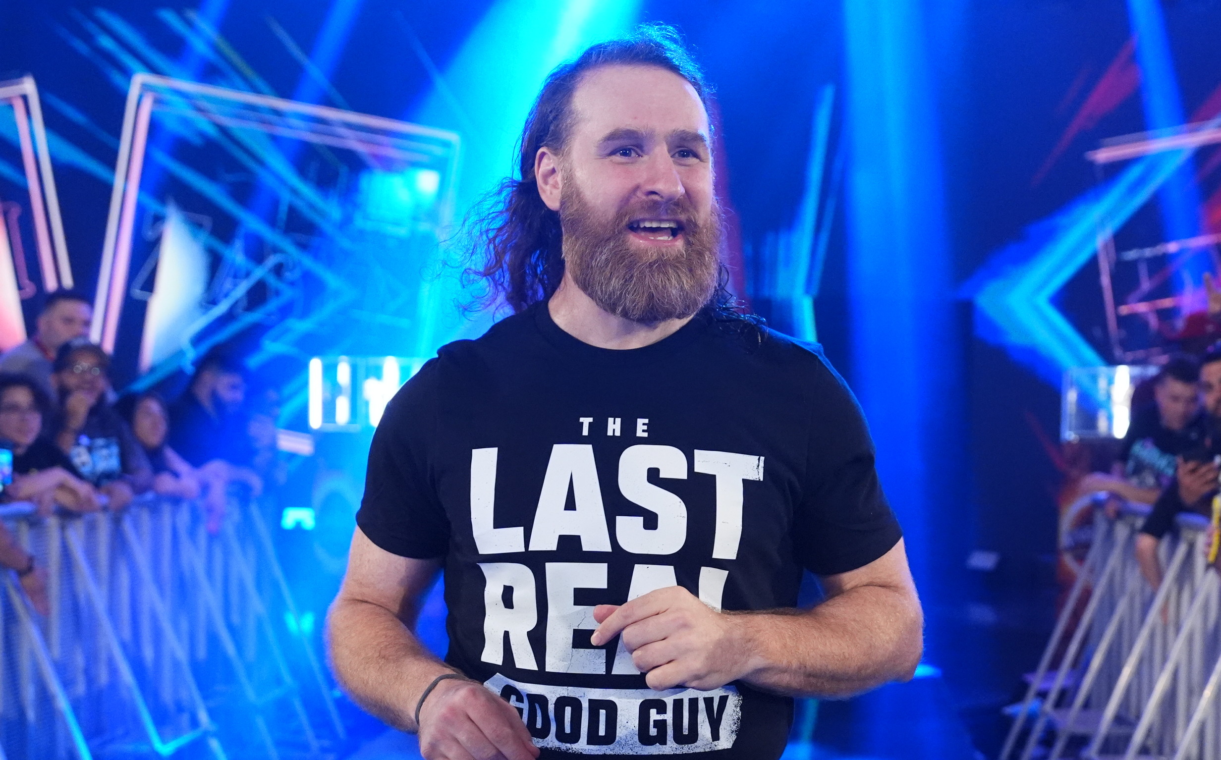 Sami Zayn with long hair and a beard walks down a lit aisle, wearing a shirt that says "The Last Real Good Guy," surrounded by cheering fans.