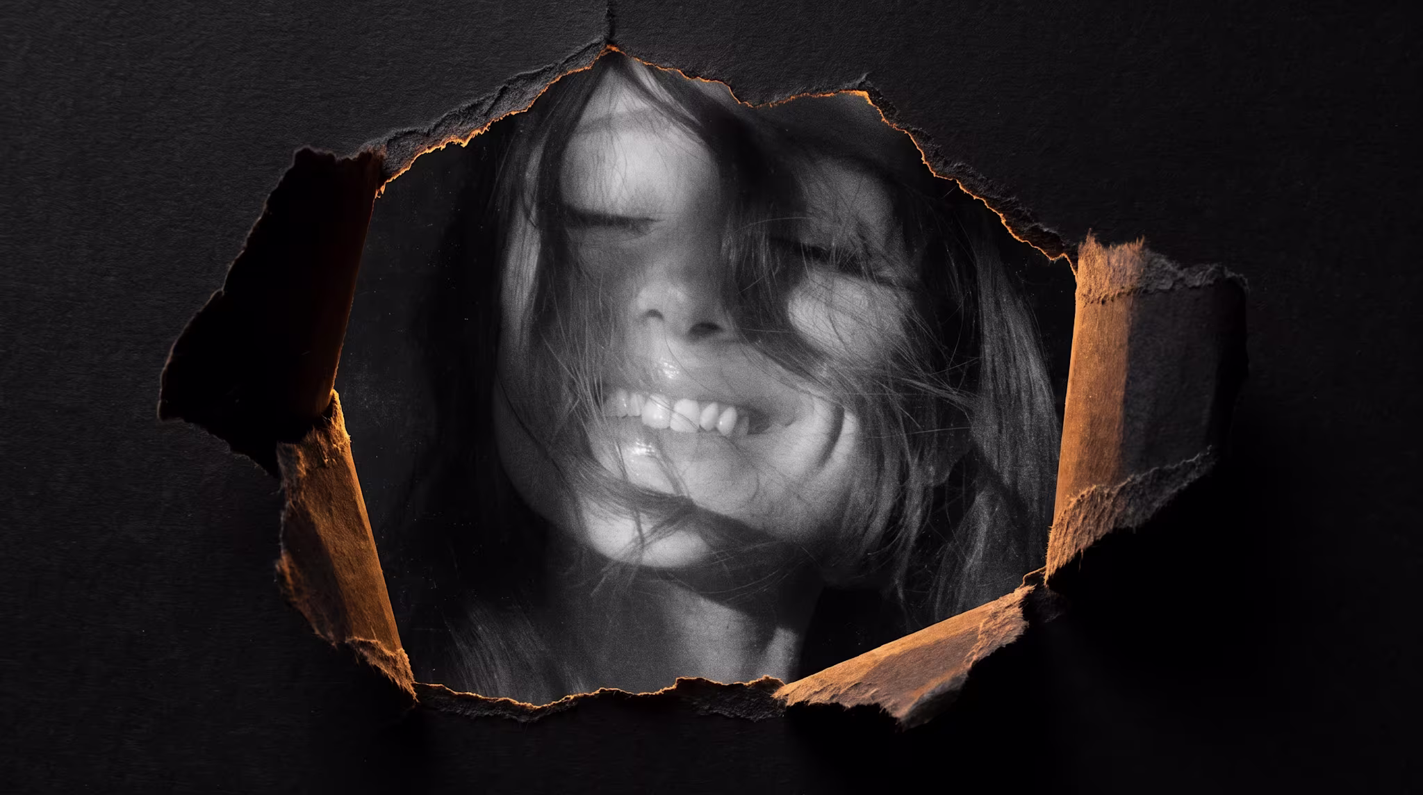 A black-and-white photo of Ariana Grande smiling with hair covering part of her face, seen through a torn paper opening on a dark background.