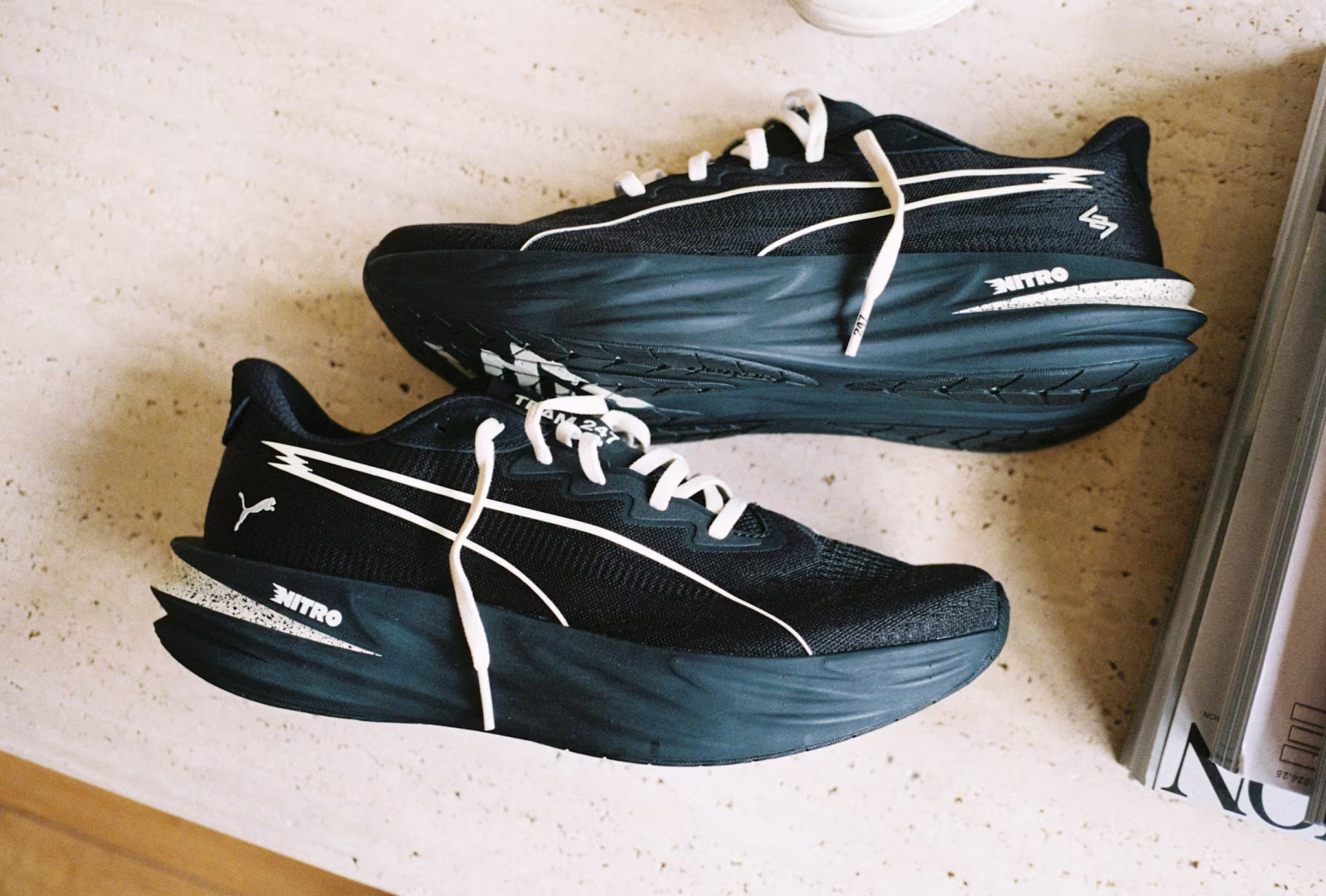 A pair of black and white Puma running shoes with thick soles on a light surface, next to a magazine.