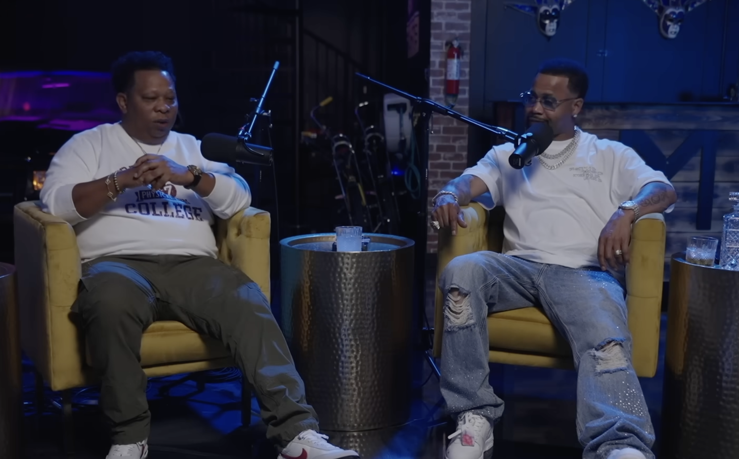 Two men sitting in chairs, Mannie Fresh and Juvenile, engaged in conversation on a podcast set with microphones and drinks on a table between them.