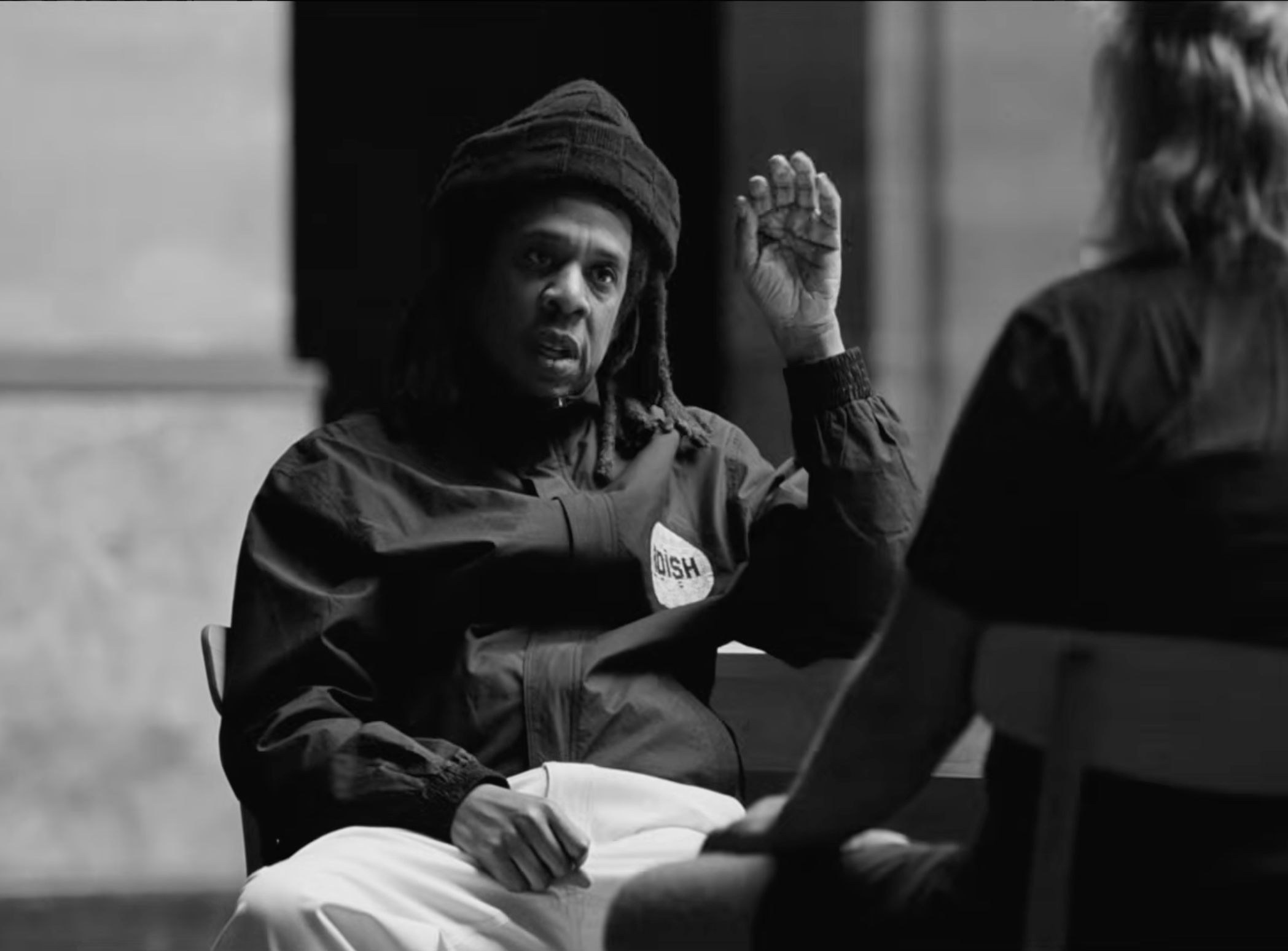 Jay-Z in a beanie and jacket gestures while sitting across from another person in a black and white setting. This is a scene from JAY-Z in 8, a docuseries from Rick Rubin.
