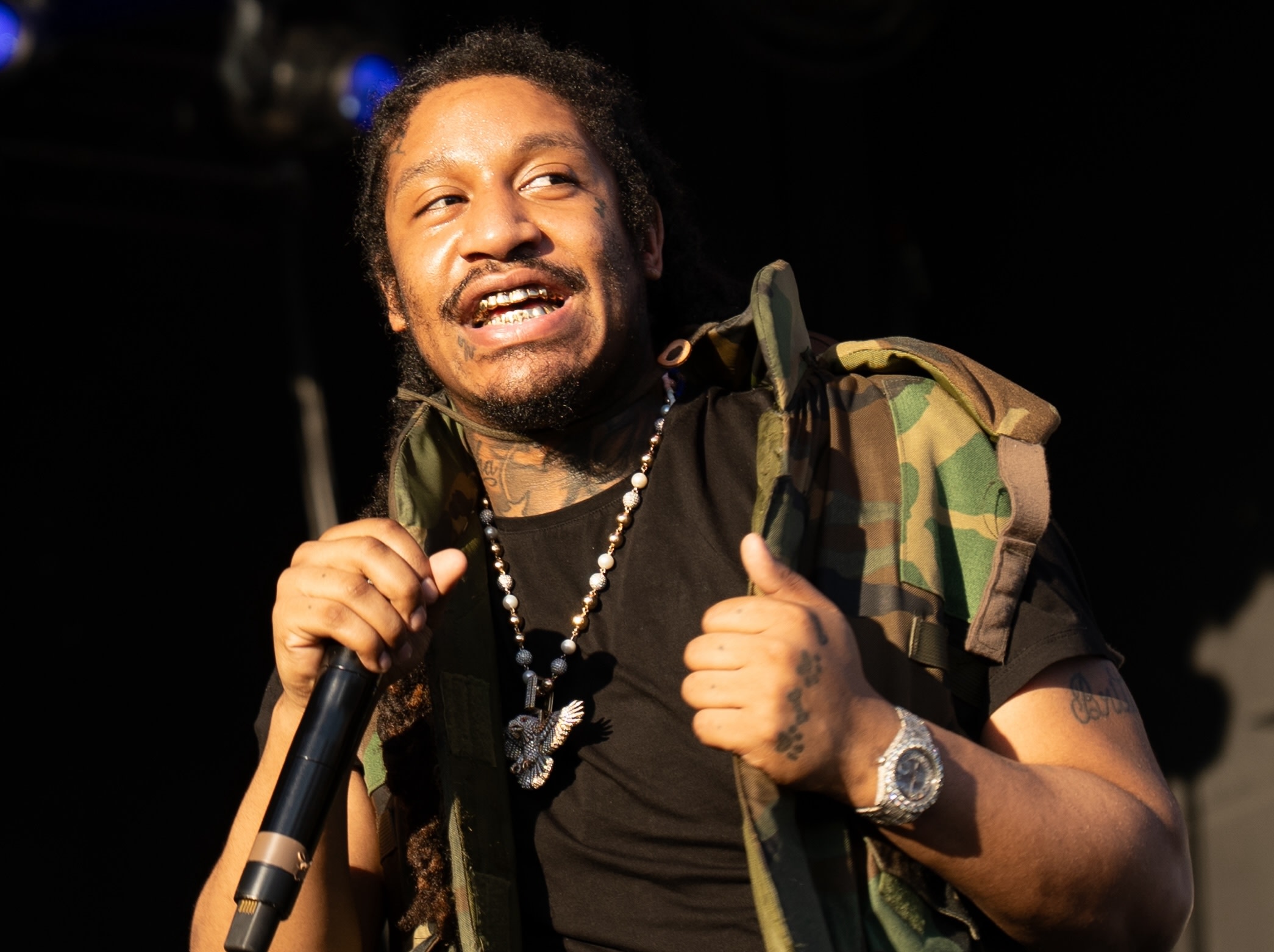 A man with braids and tattoos, wearing a camo vest and holding a microphone, smiles on stage.