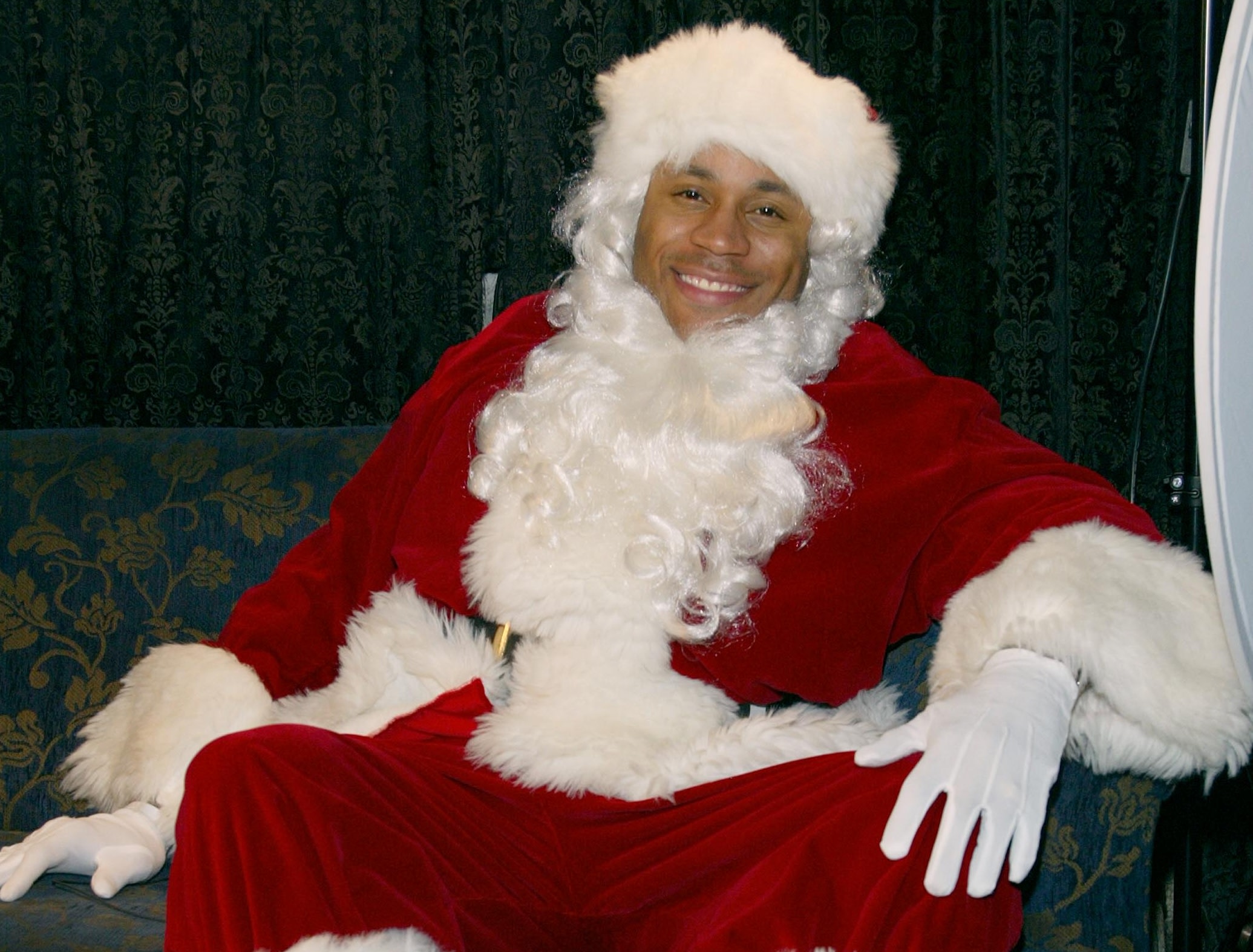 Rapper LL Cool J dressed in a red and white Santa Claus outfit, sitting on a patterned couch, smiling at the camera.