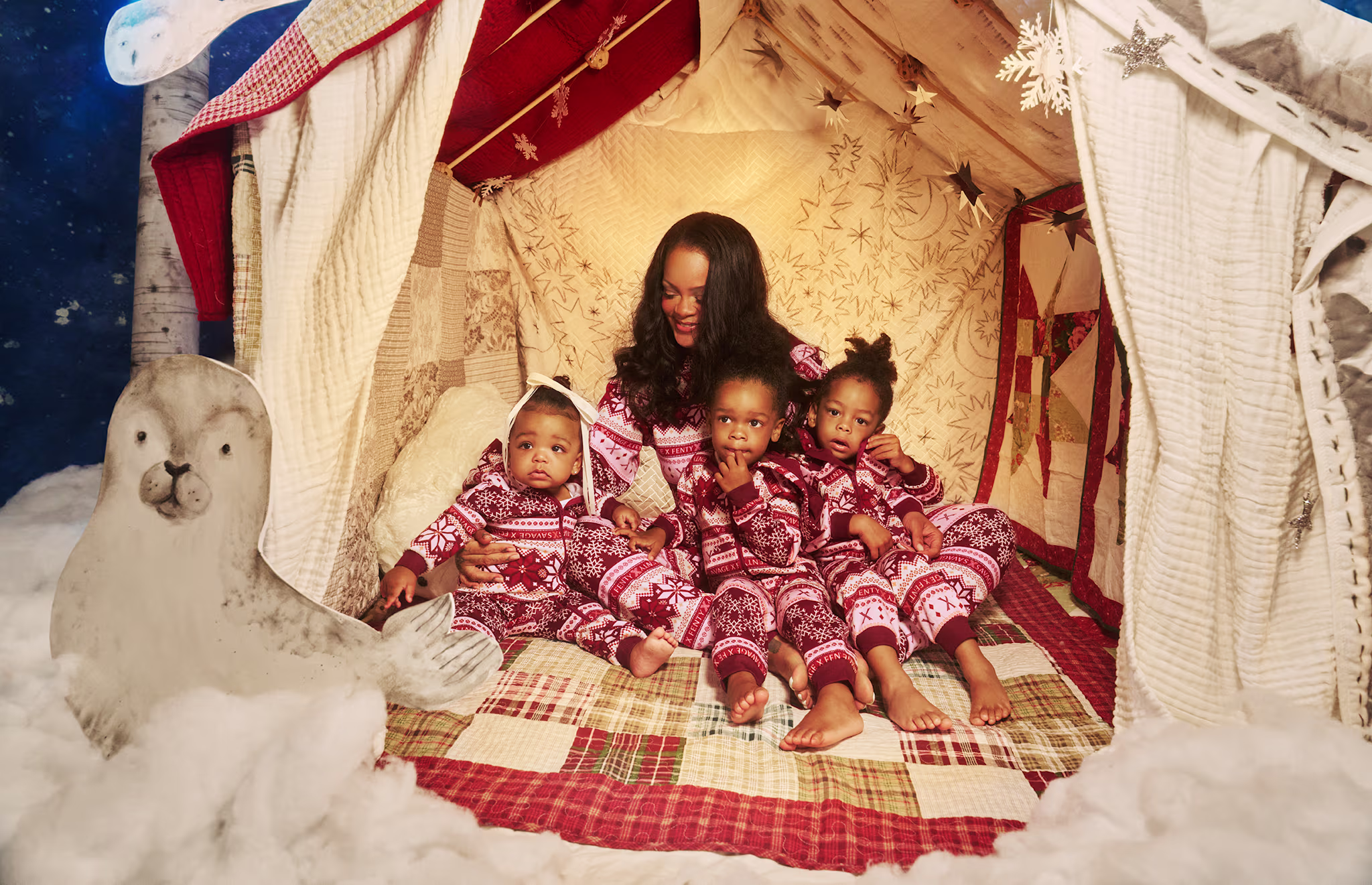 A woman and three children in matching red Christmas pajamas sit inside a cozy, decorated tent with snow and a polar bear figure outside.