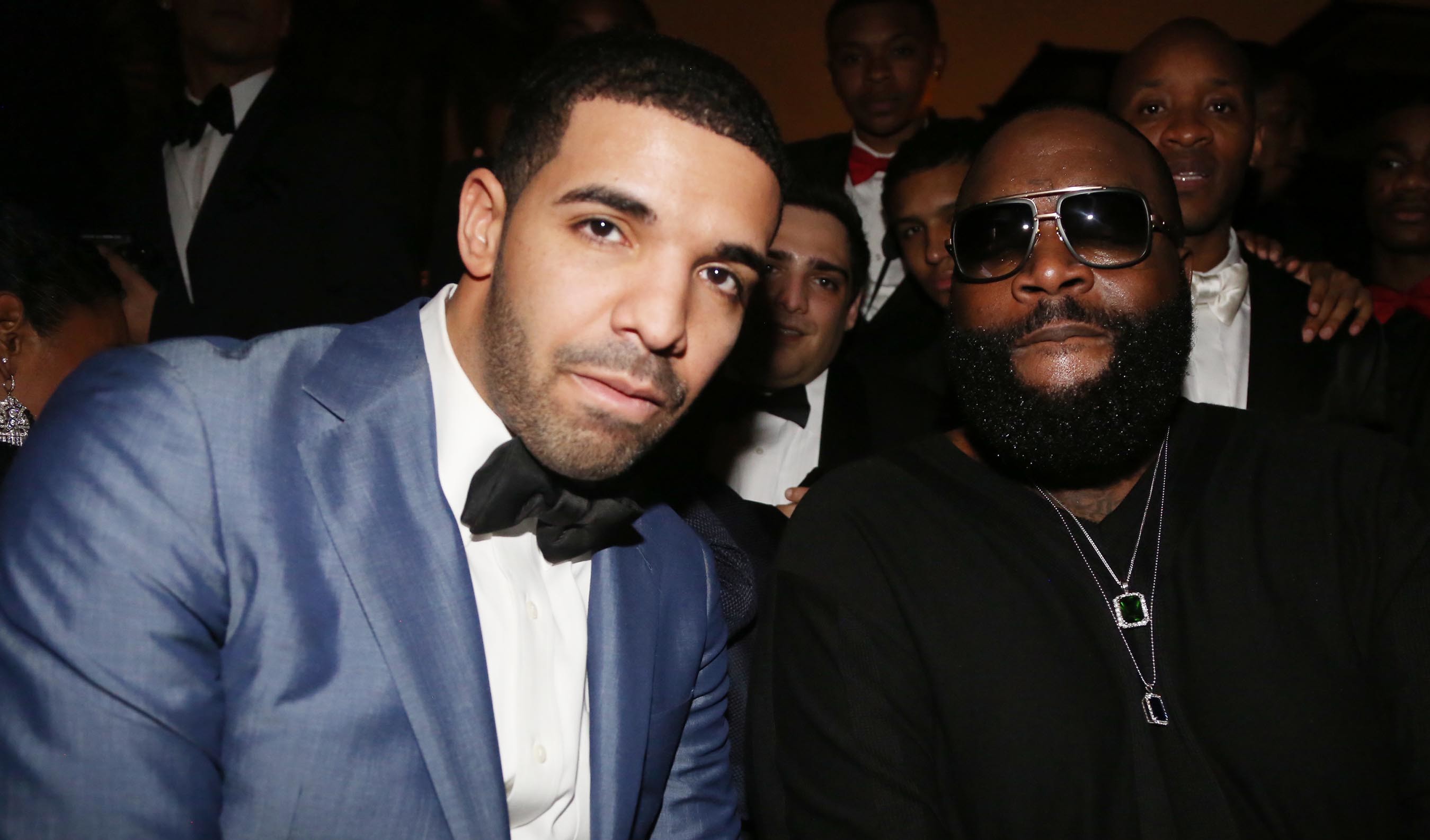 Drake in a blue tuxedo and Rick Ross in black attire with sunglasses, posing together at an event with people in the background.