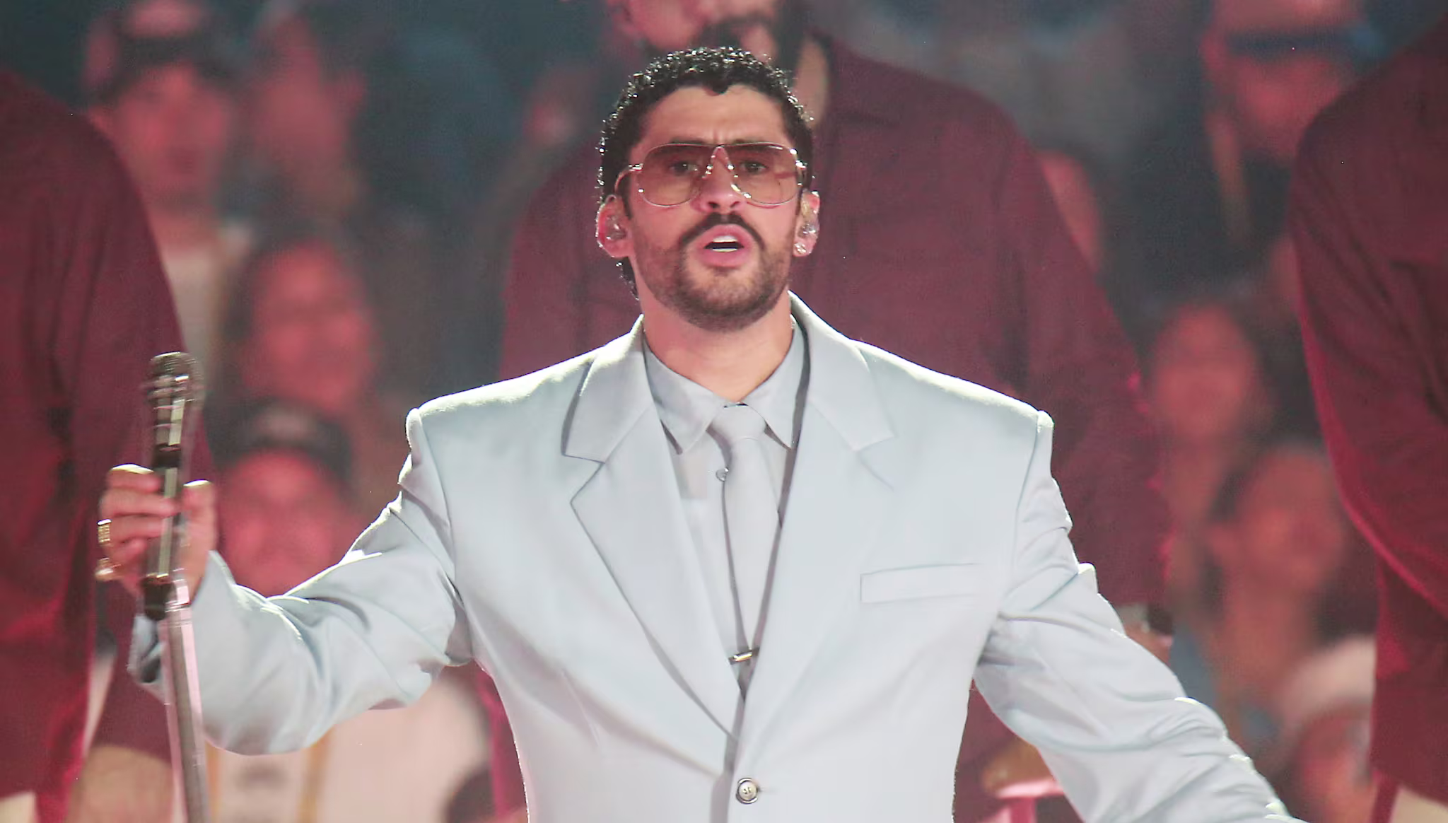 Bad Bunny in a light blue suit performs on stage with backup dancers in red shirts and hats, in front of a cheering crowd.