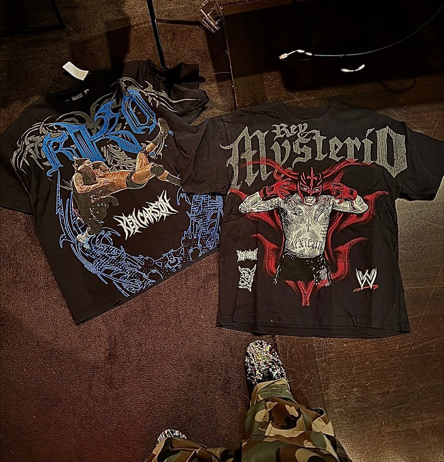 Two WWE t-shirts on the floor: one featuring Rey Mysterio, the other with an RKO design. Person in camo pants and patterned shoes.