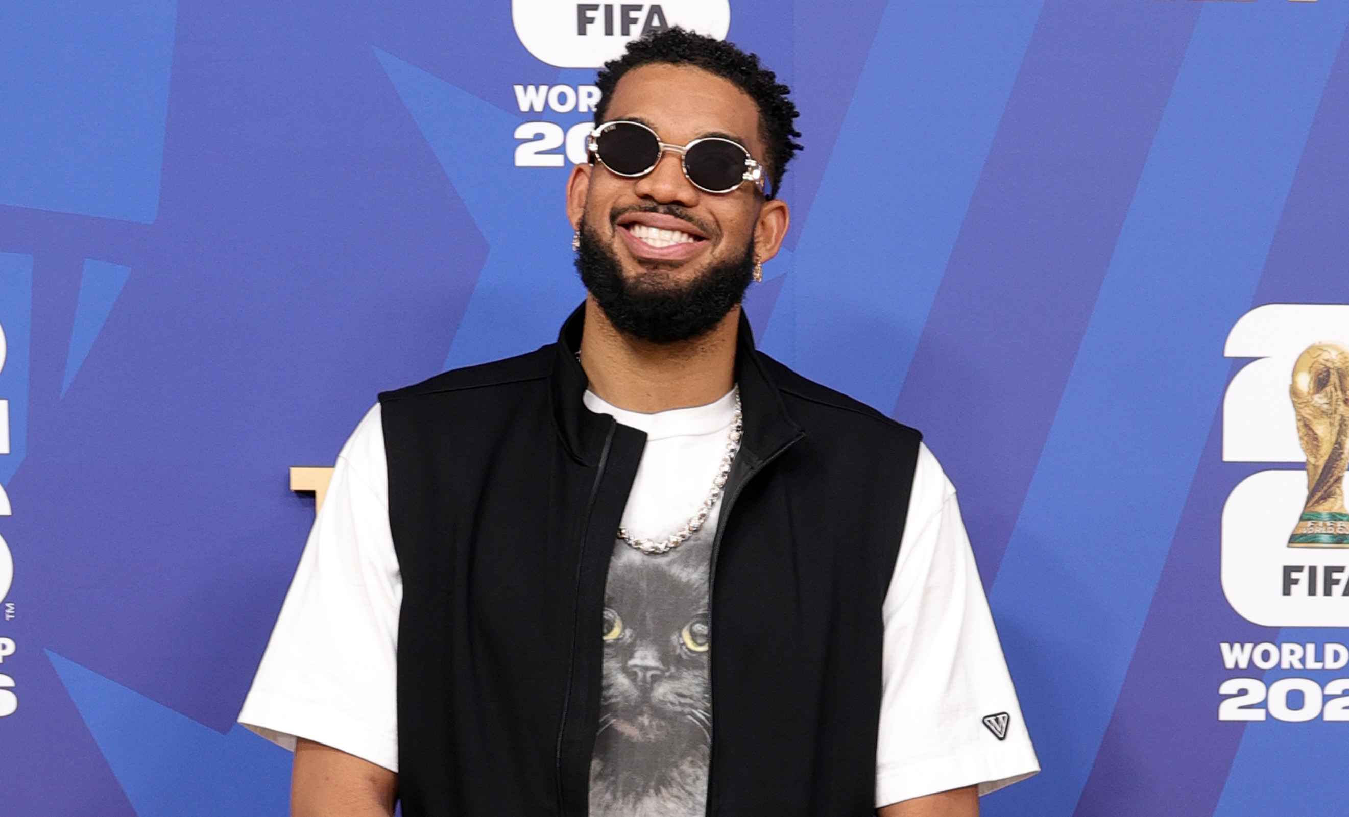 Karl-Anthony Towns in sunglasses, a black vest, white t-shirt, and baggy jeans with patches, stands smiling in front of a FIFA World Cup 2026 backdrop.