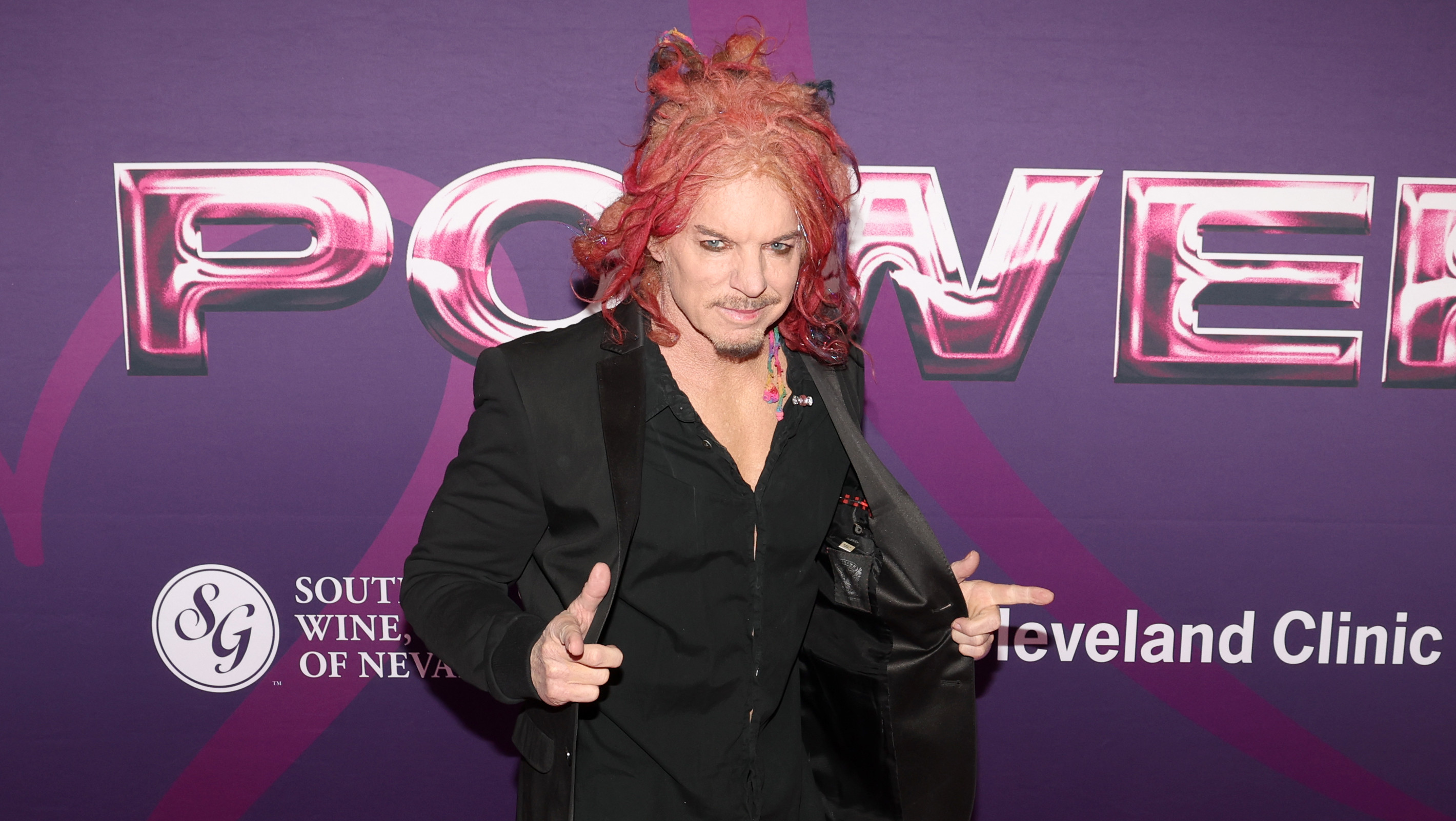 Carrot Top with red hair stands on a pink carpet at an event. The backdrop features logos and the words "Power of Love."