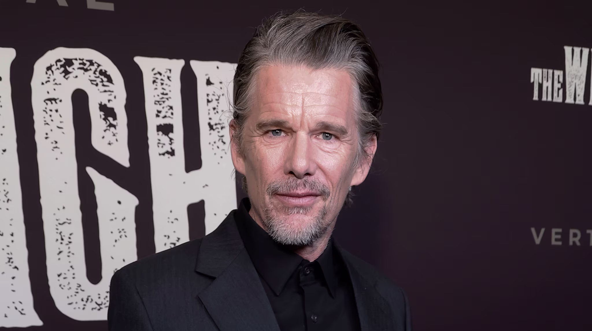 Ethan Hawke in a dark suit at an event, holding sunglasses, with a microphone and paper visible in the foreground.