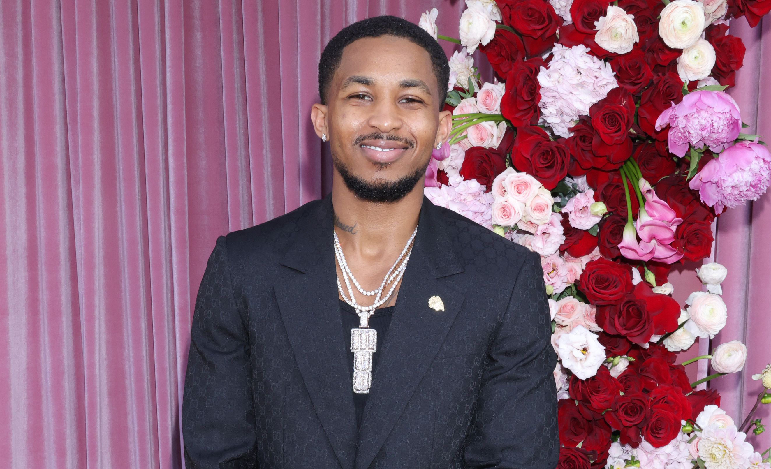 DDG in a black suit stands on a red carpet with a floral backdrop.