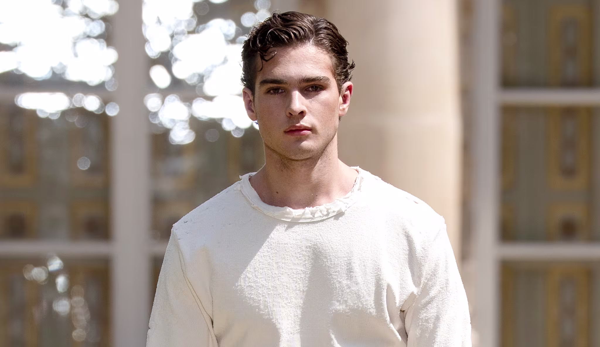 Clavicular in a white outfit walking outdoors, with a neutral expression and styled hair.