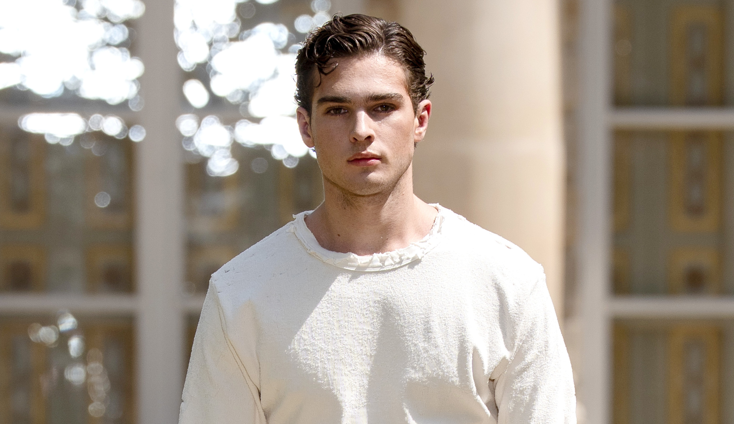 Clavicular in a white outfit walking outdoors, with a neutral expression and styled hair.