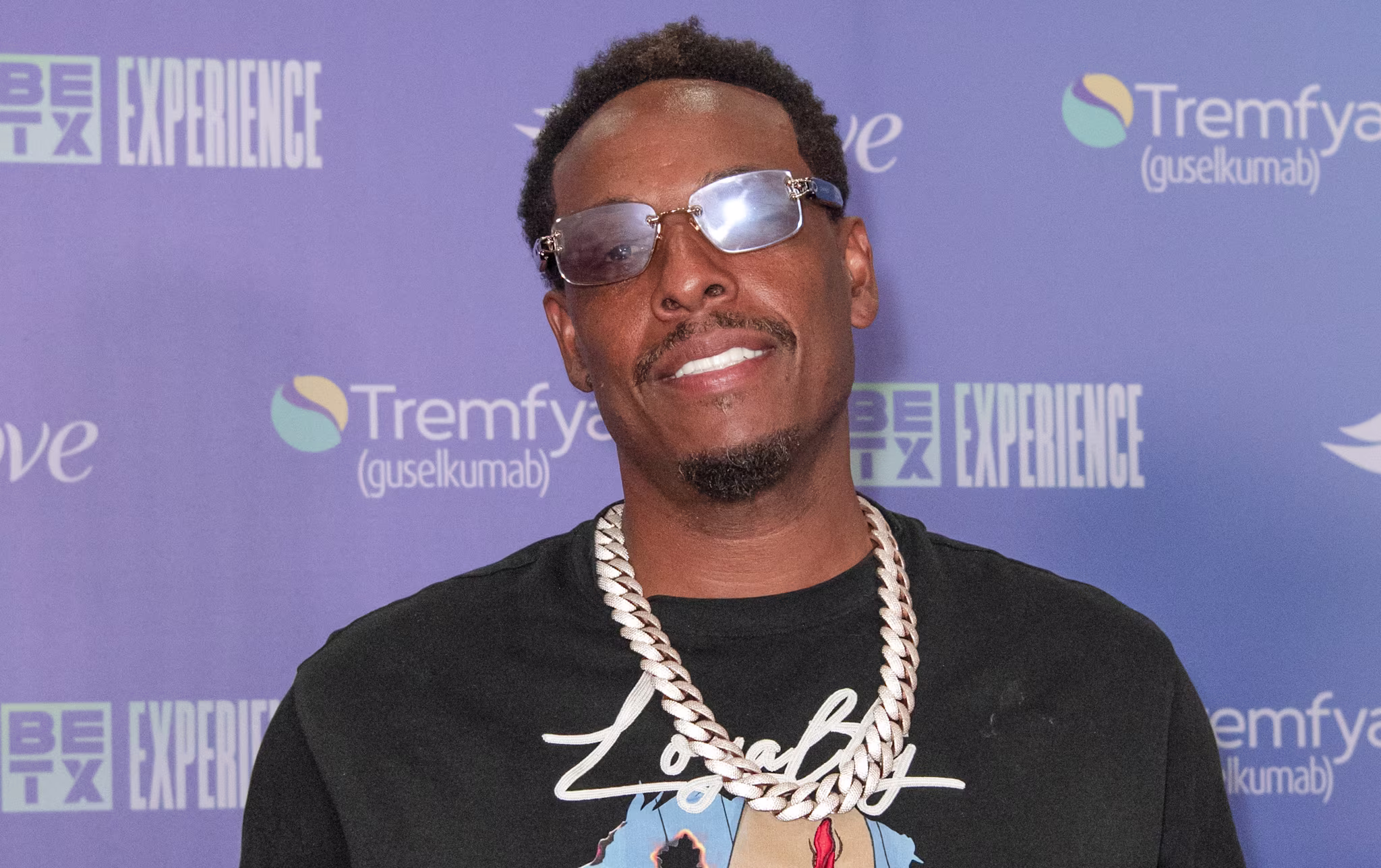 Paul Pierce in sunglasses and a graphic t-shirt poses with thumbs up at a BET Experience event backdrop.