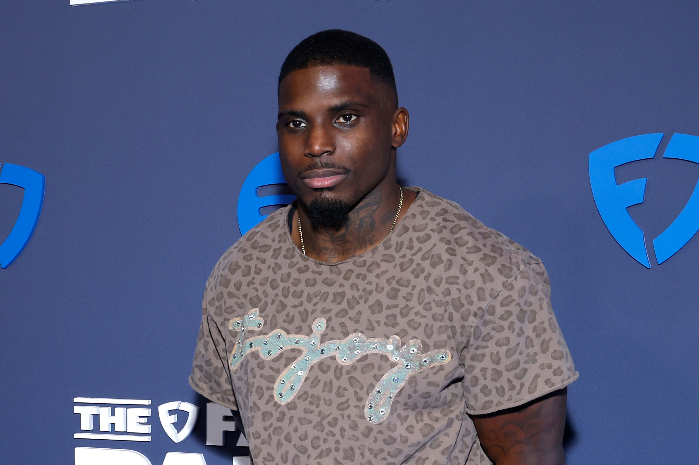 Tyreek Hill stands on a blue carpet at a FanDuel event, wearing a patterned shirt and striped pants, with a blue backdrop featuring logos.