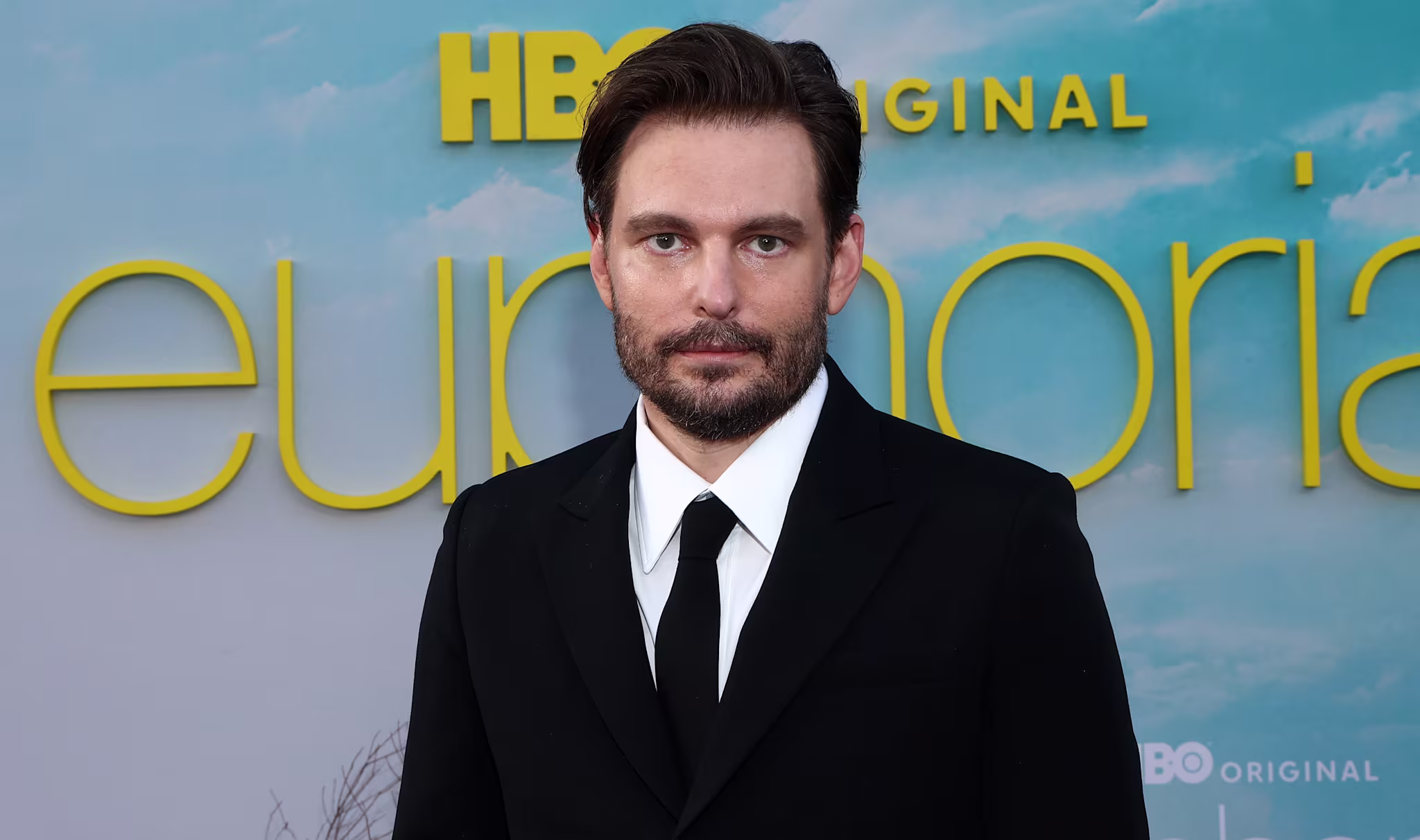 Sam Levinson in a black suit stands in front of a "Euphoria" backdrop with the HBO logo.