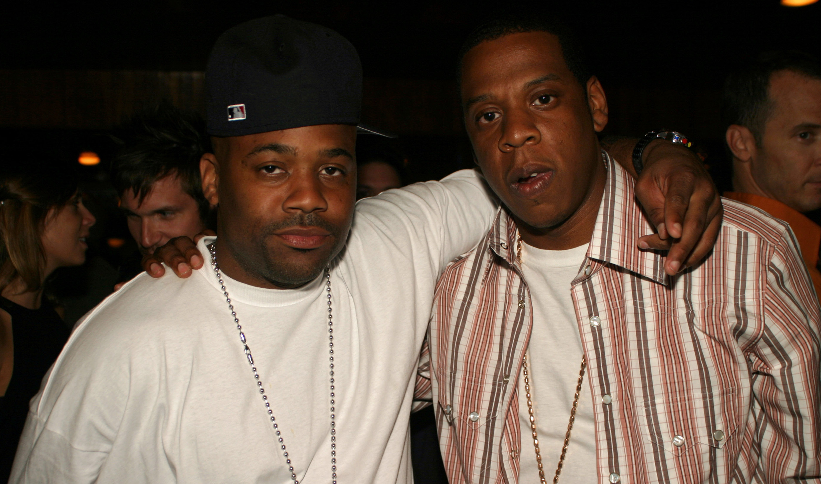 Damon Dash and Jay-Z posing together, both wearing necklaces. Jay-Z is in a striped shirt, and Damon Dash in a white shirt and cap.