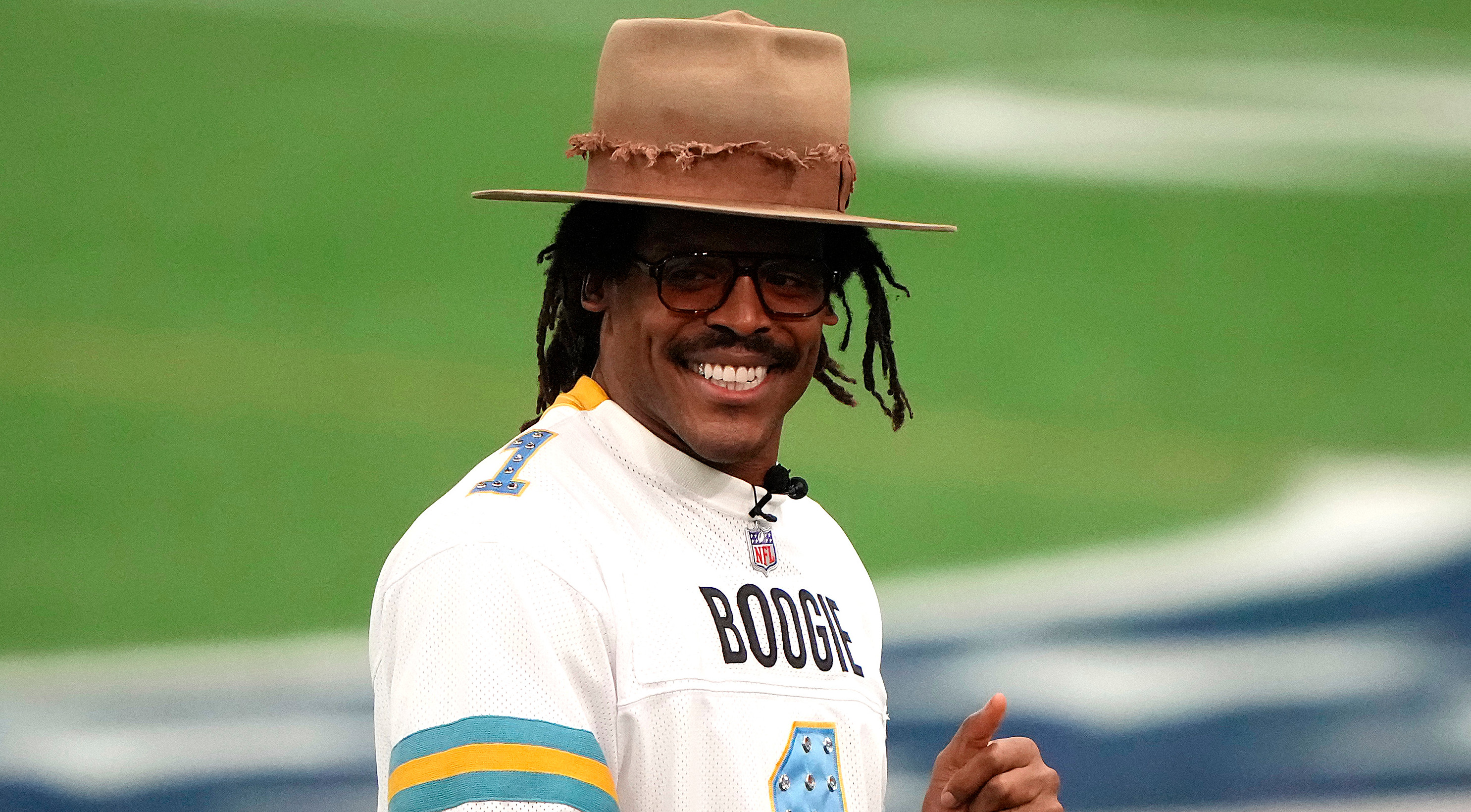 Cam Newton wearing a wide-brimmed hat, glasses, and a football jersey with "Boogie" on it, smiling and giving a thumbs-up on a field.
