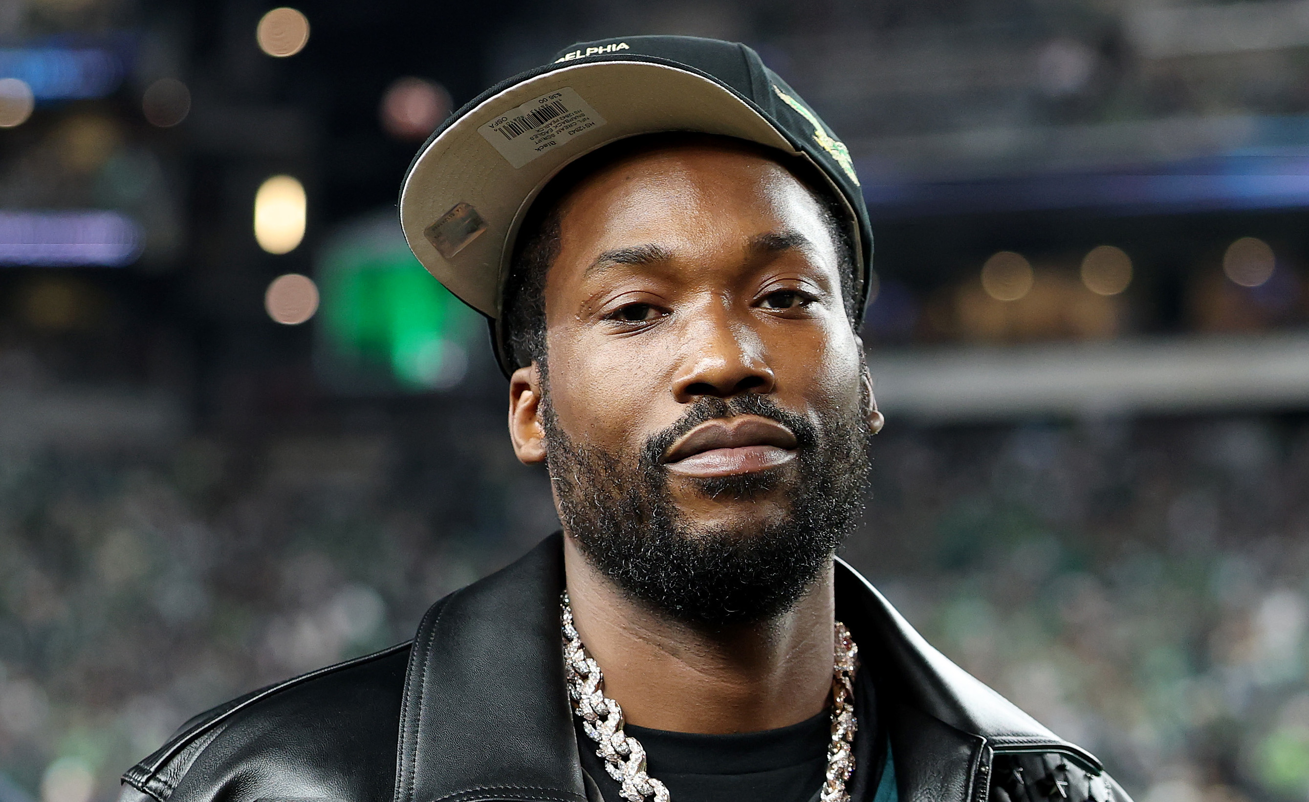 Meek Mill wearing a Philadelphia Eagles jacket and cap, with a large chain necklace, standing in a stadium.