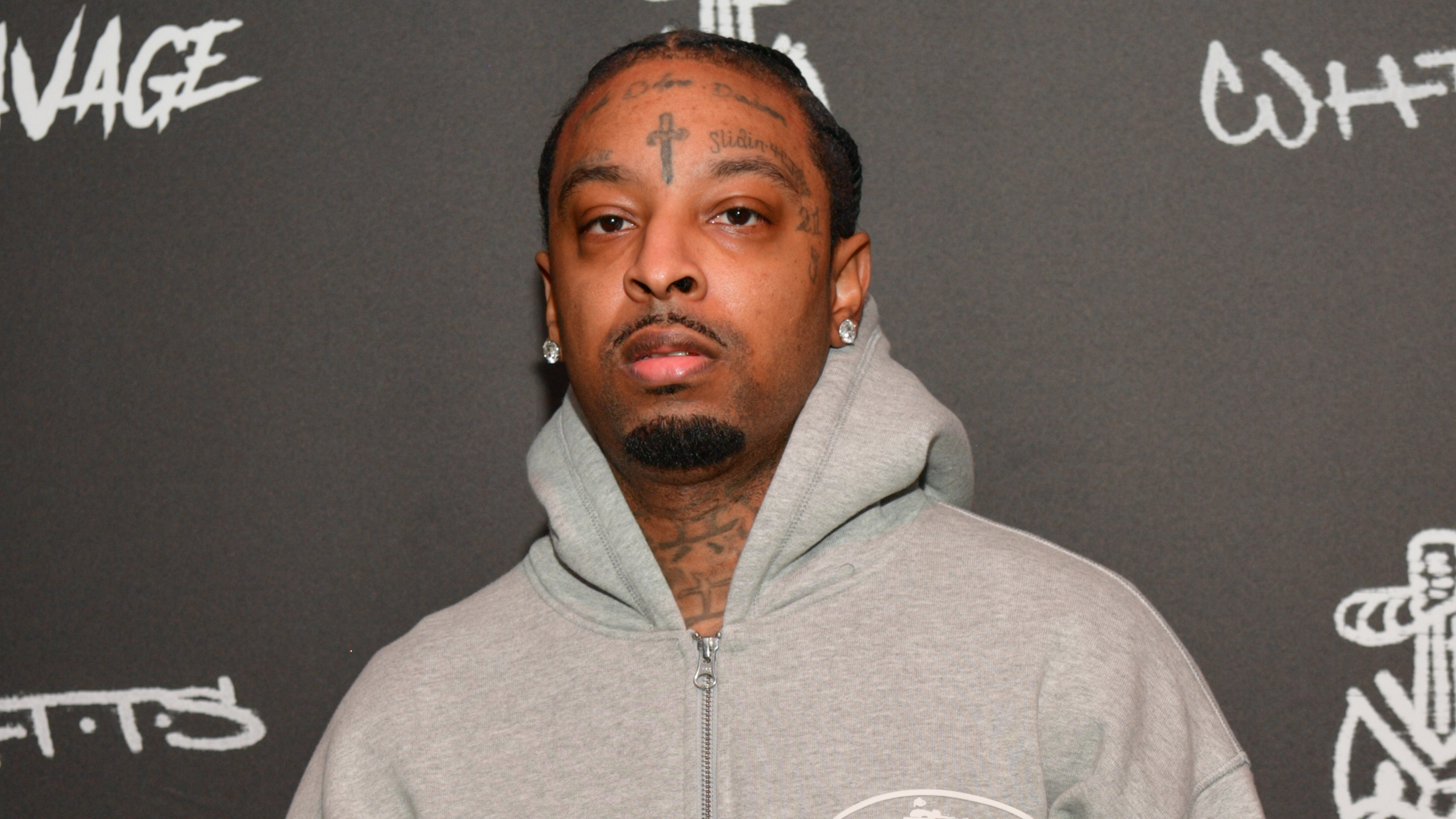 Rapper 21 Savage in a gray hoodie, standing against a dark background with graffiti-style text.