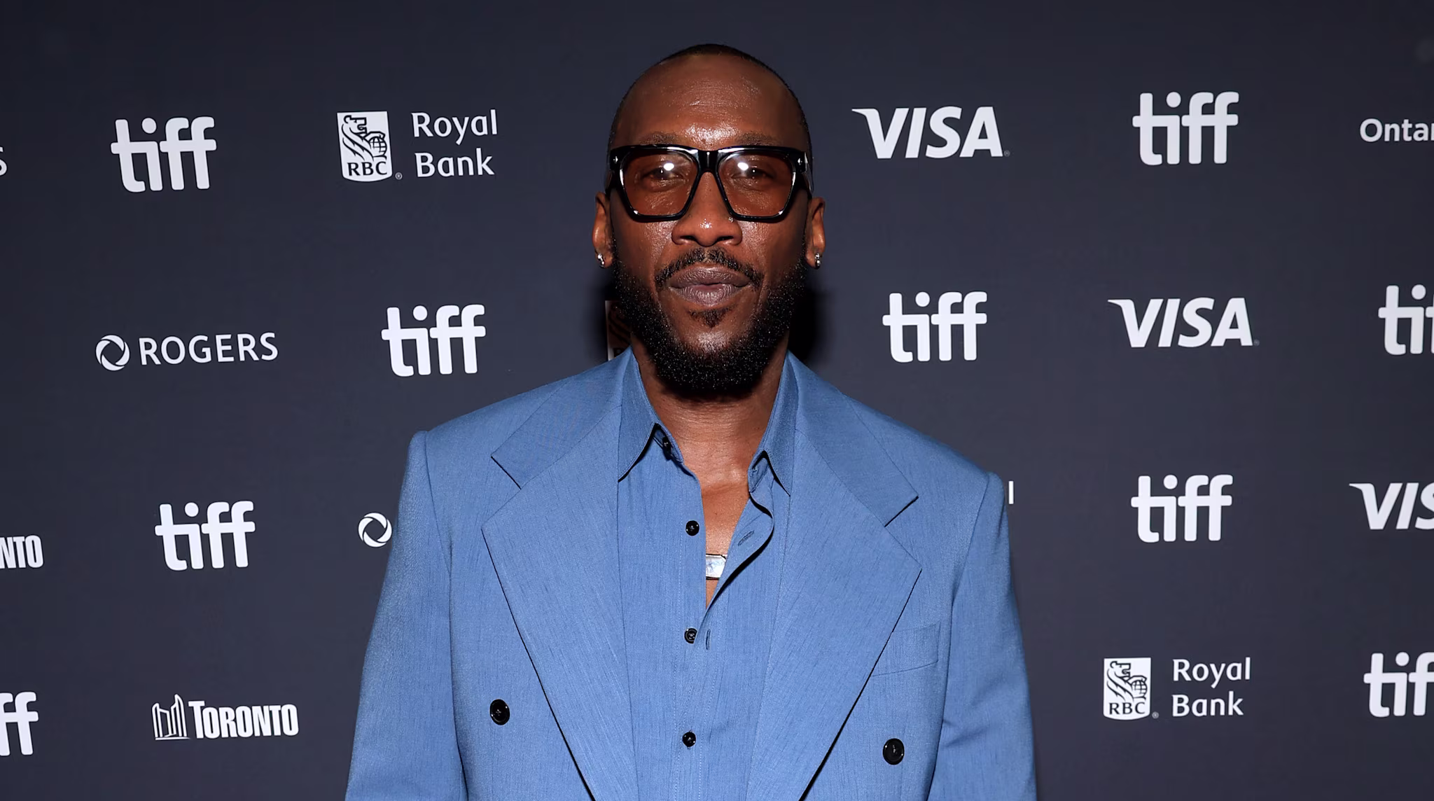 Mahershala Ali in a blue suit and glasses stands on the red carpet at a TIFF event, with a branded backdrop behind him.