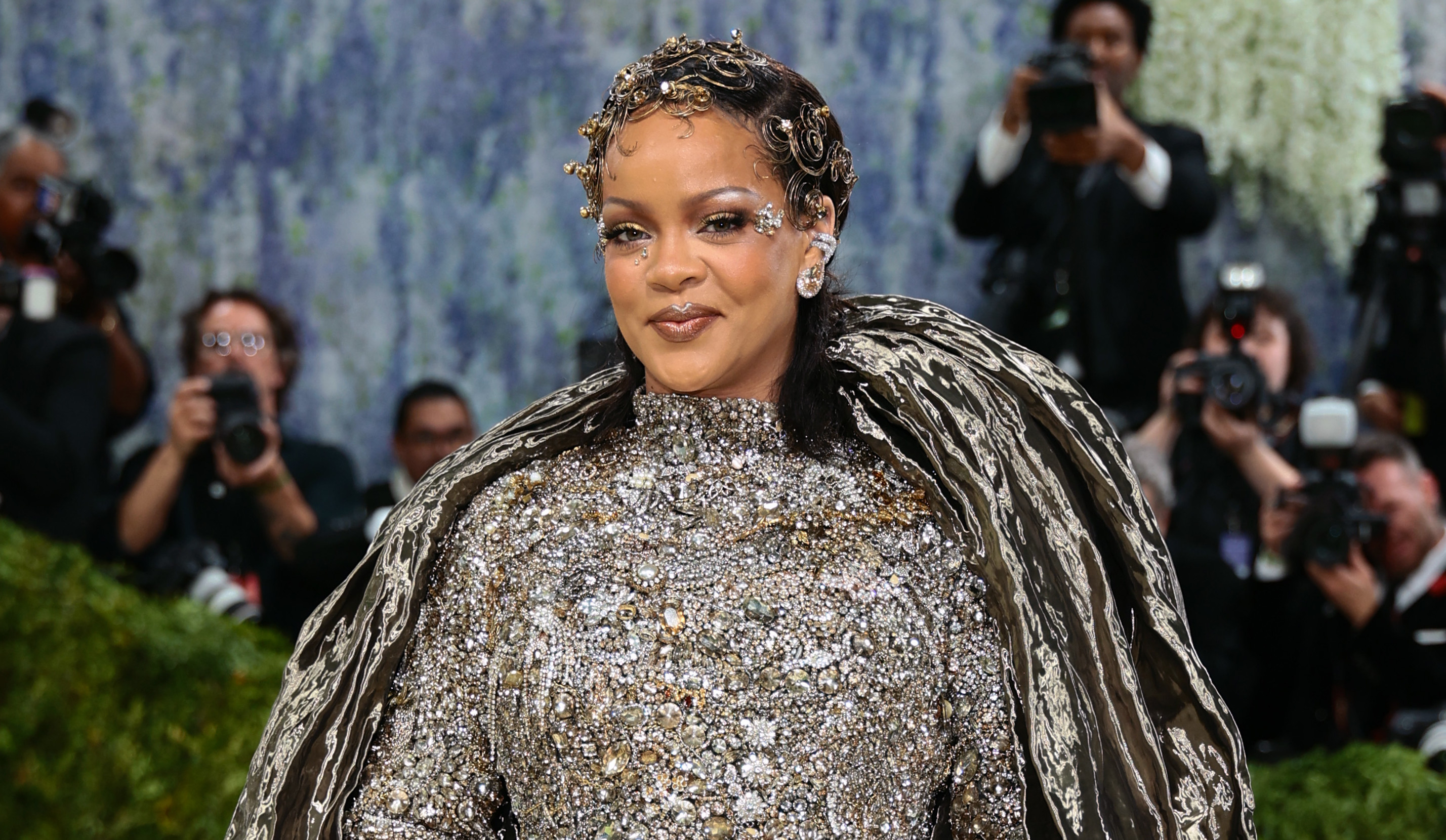 Rihanna at a fashion event, wearing a shimmering silver gown with a dramatic circular design, surrounded by photographers.