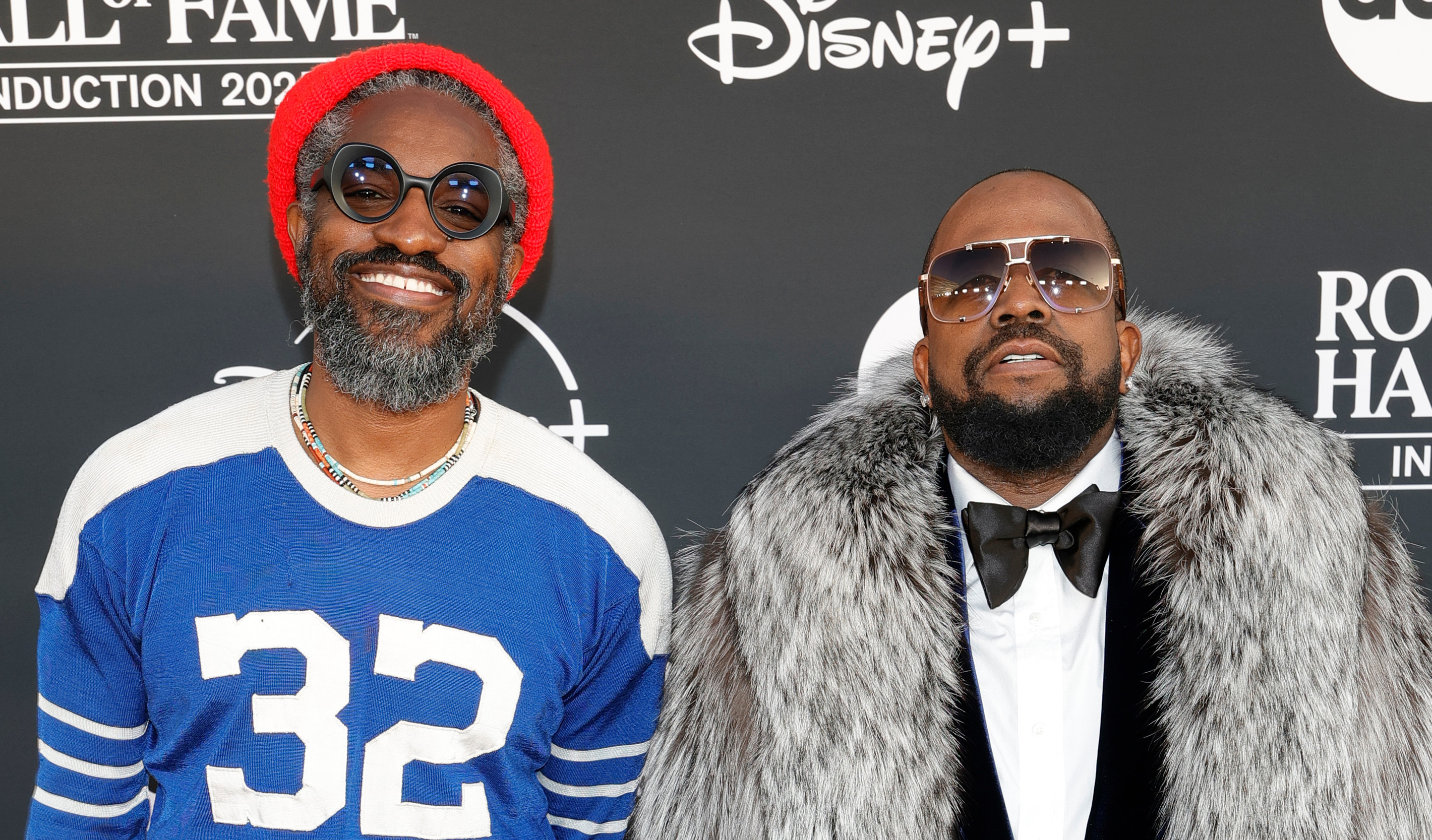 OutKast on the red carpet at the Rock & Roll Hall of Fame event. Andre 3000 wears a blue jersey and red hat, Big Boi in a fur coat and sunglasses.