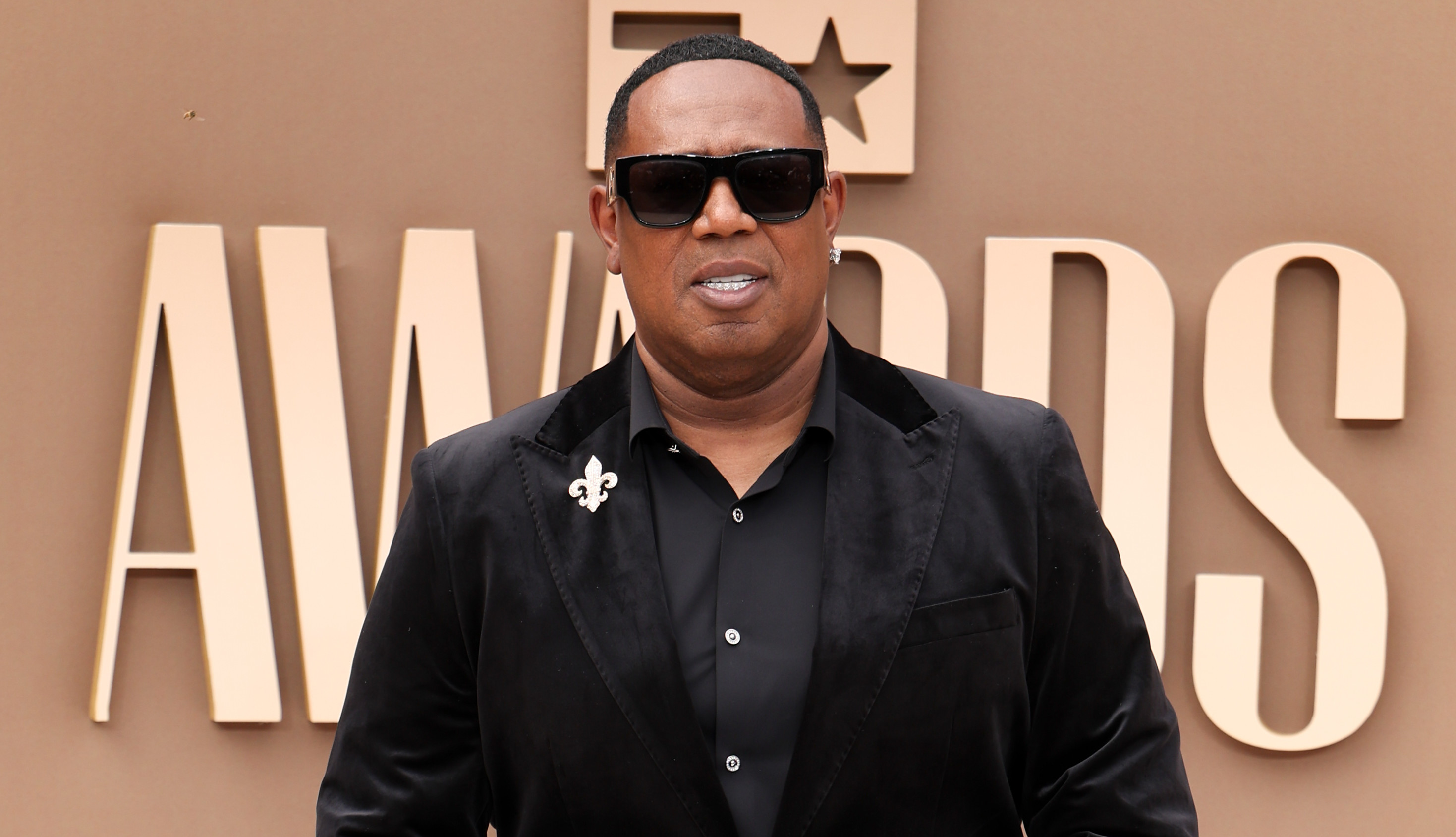 Master P in black jacket and checkered pants at an awards event, wearing sunglasses and jewelry, standing on a circular carpet.