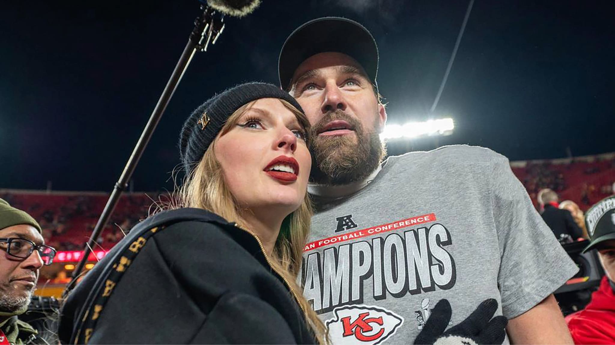 Taylor Swift and Travis Kelce in a Kansas City Chiefs "Champions" shirt at a stadium event.