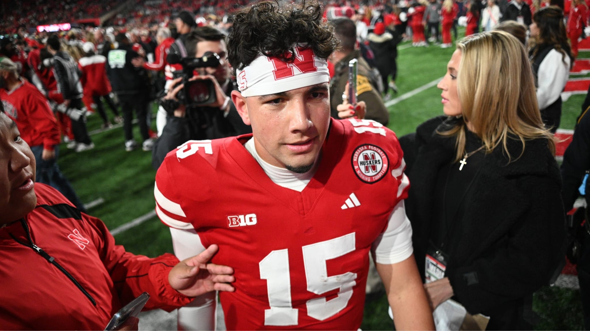 Dylan Raiola in a red Nebraska uniform with number 15 on the field, surrounded by people and media.