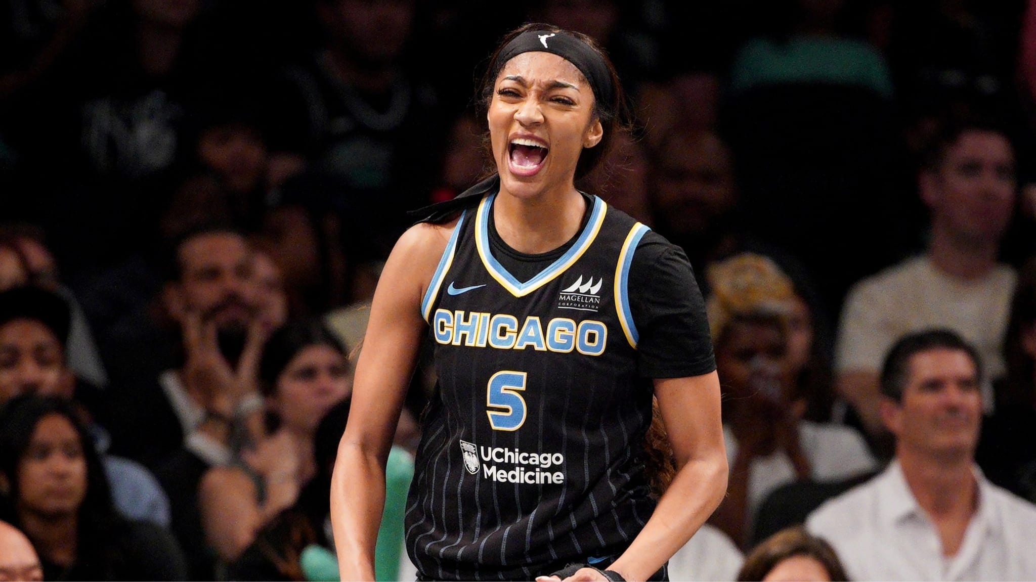 Angel Reese in a Chicago uniform, number 5, is energetically celebrating on the court with a cheering crowd behind her.