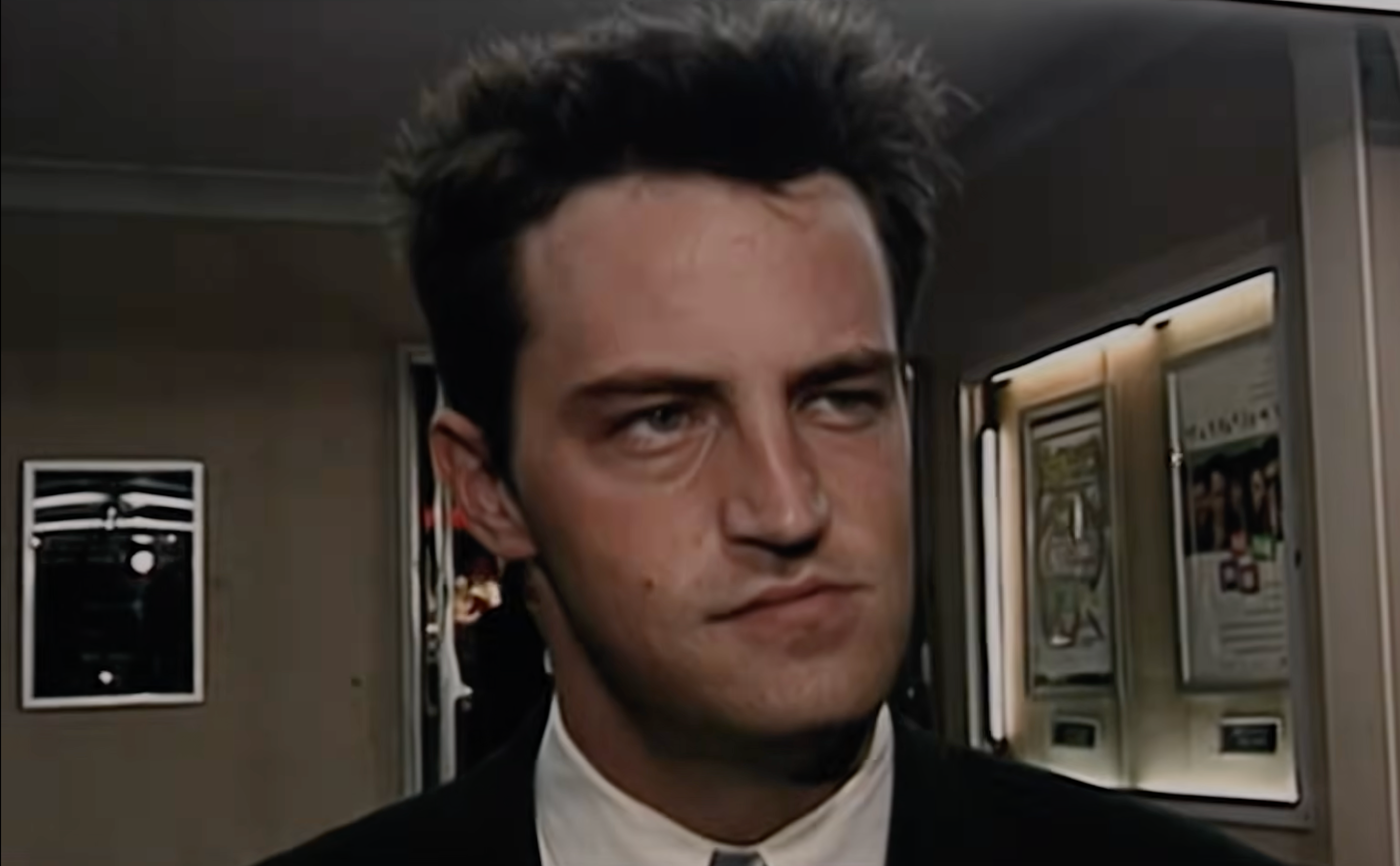 Matthew Perry with dark hair in a suit stands indoors, looking to the side. The background shows framed posters on the wall.