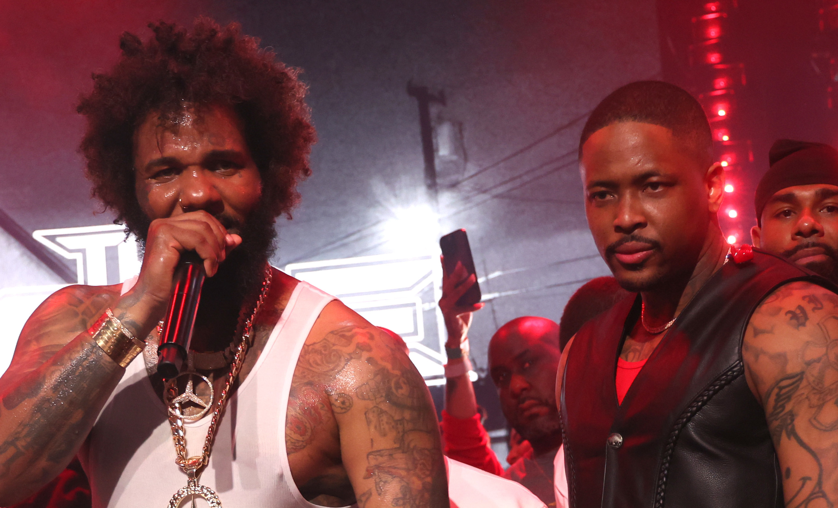 Rappers The Game and YG performing on stage, surrounded by a crowd, with red lighting.