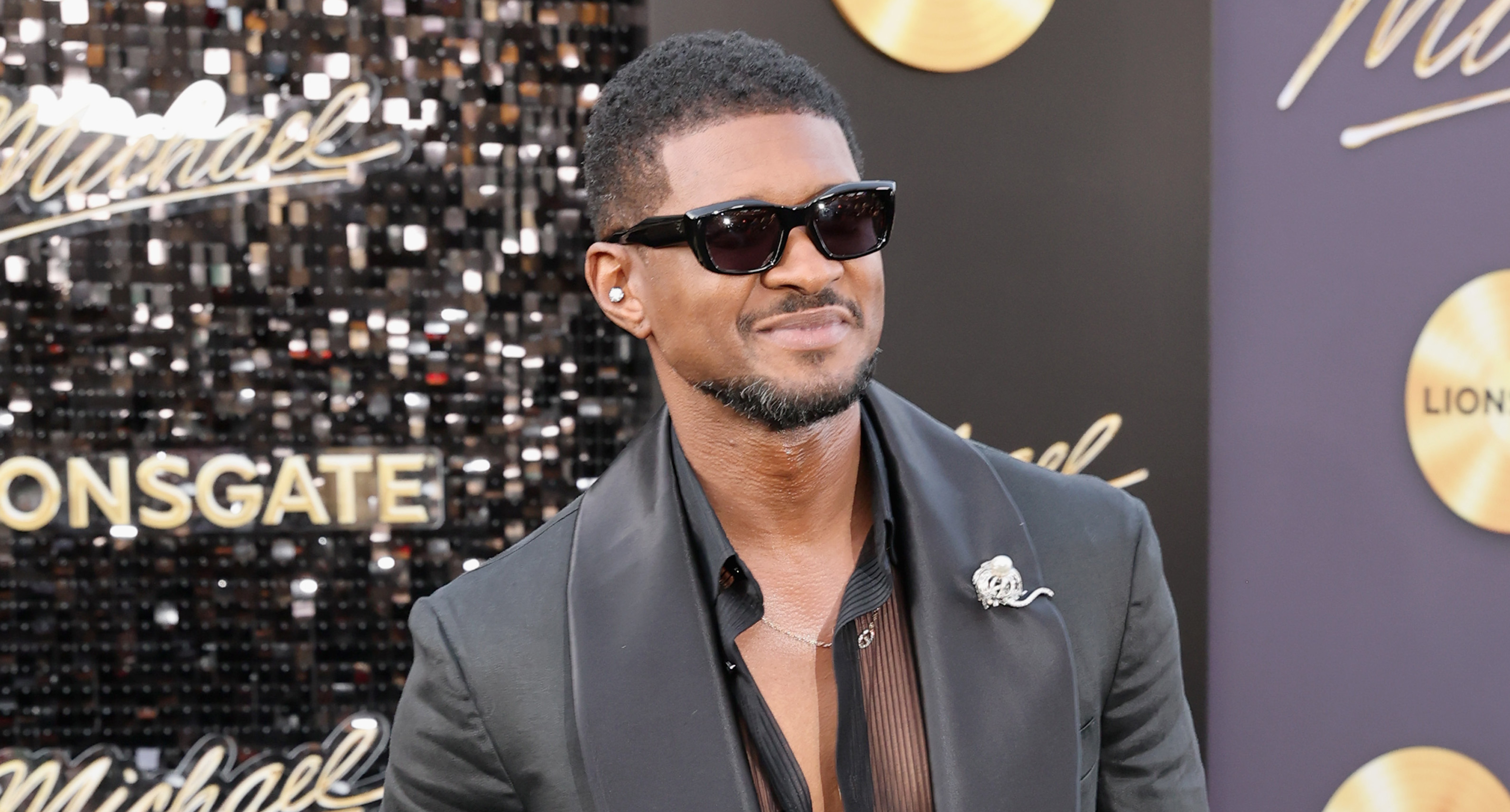 Usher in a stylish black suit with sunglasses, standing on a red carpet at a Lionsgate event.