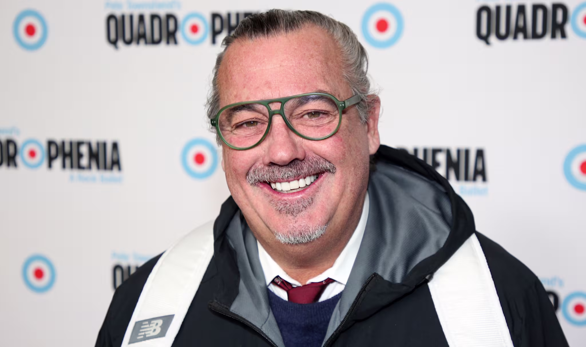Duncan Sheik wearing glasses, a black New Balance jacket, and a white backpack, smiling at the "Quadrophenia" event backdrop.