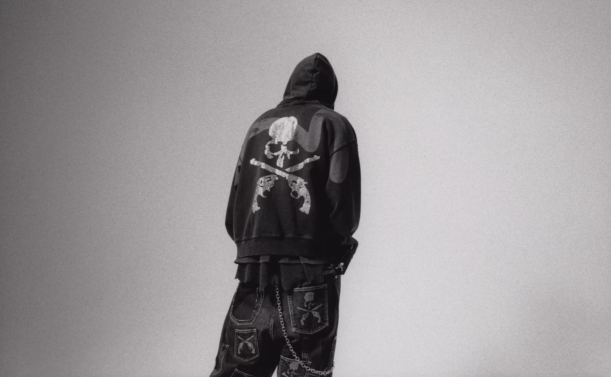 A person in a hooded jacket with a skull and crossbones design, wearing baggy pants with patches and chains, facing away.