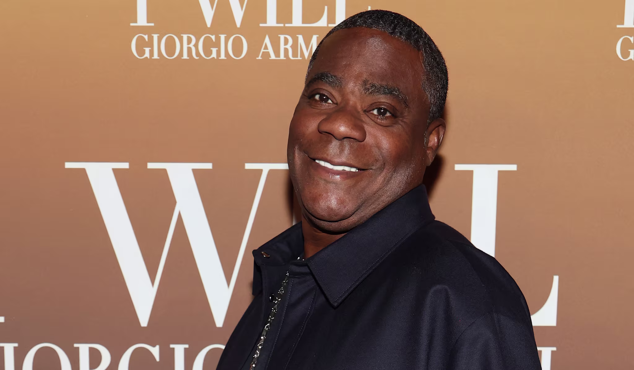 Tracy Morgan in a dark shirt poses with a smile on a Giorgio Armani backdrop.