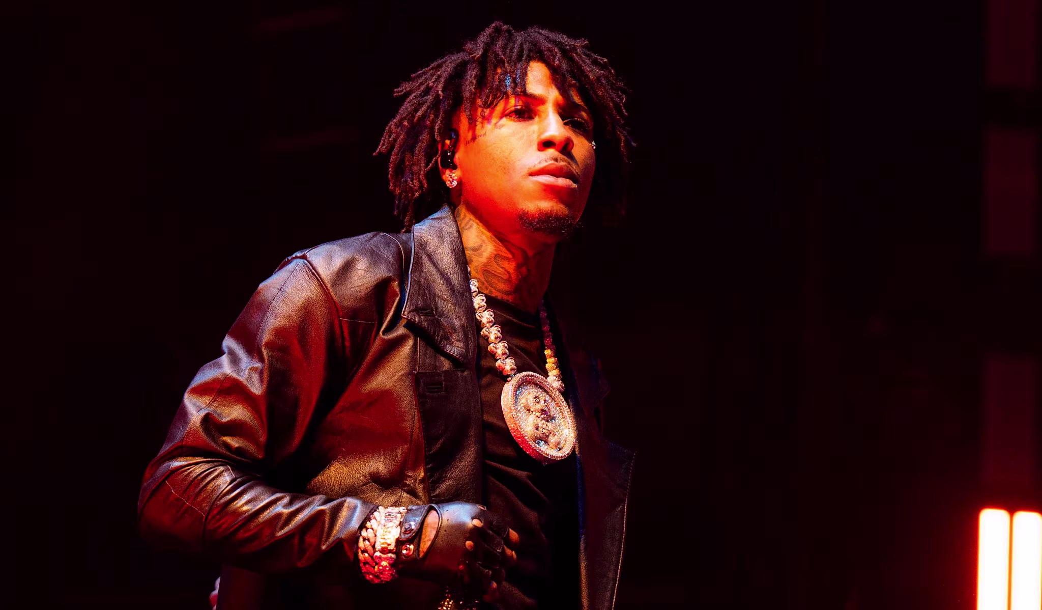 NBA YoungBoy on stage wearing a long black leather coat, with a large pendant necklace, under dramatic lighting.