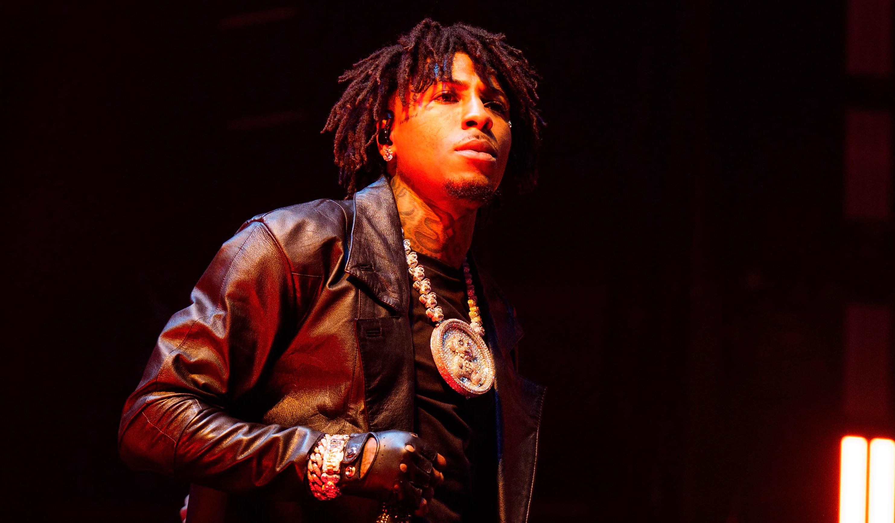NBA YoungBoy on stage wearing a long black leather coat, with a large pendant necklace, under dramatic lighting.