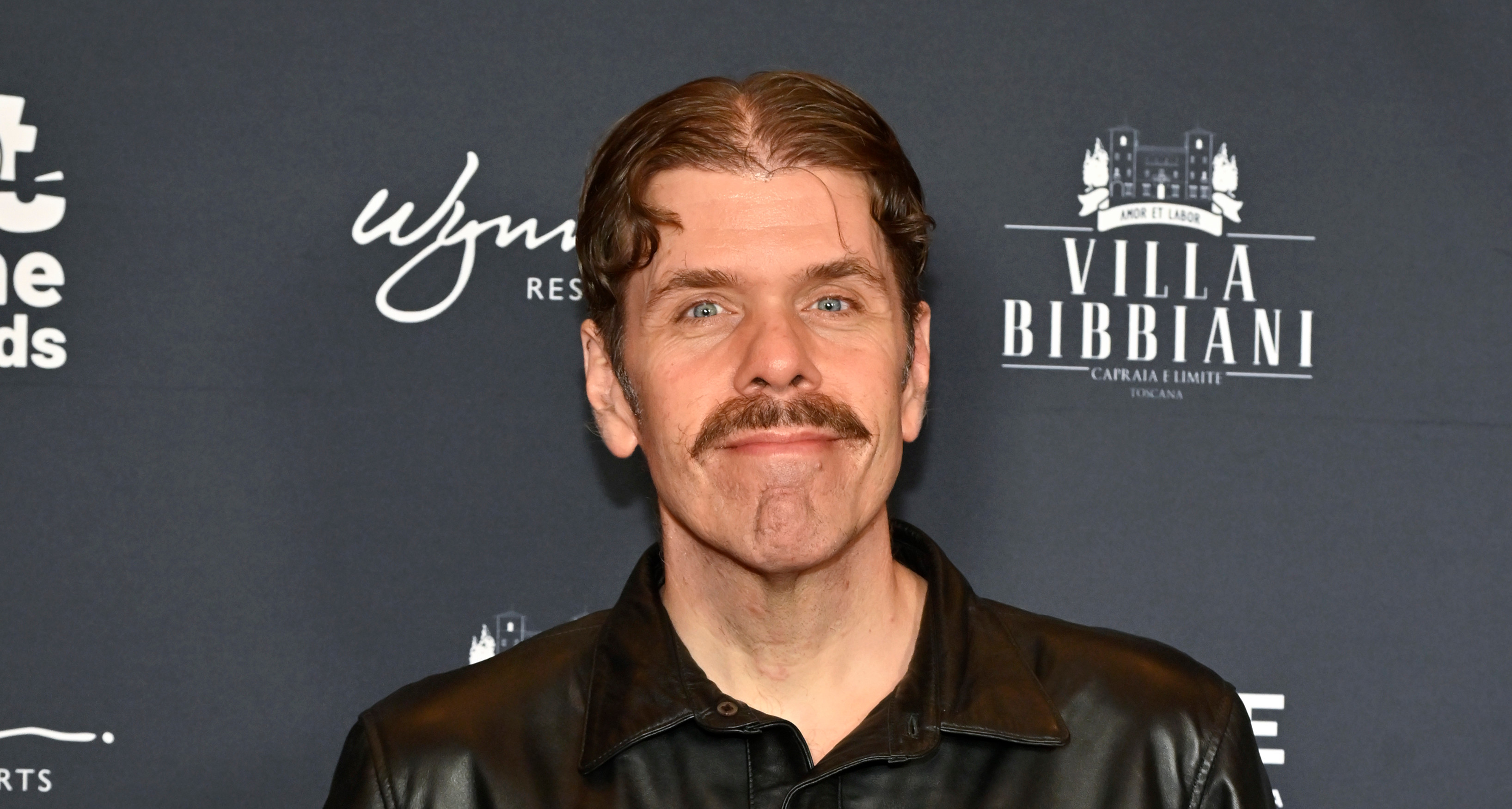 A person in a black leather jacket poses on a red carpet with a dark backdrop featuring logos.
