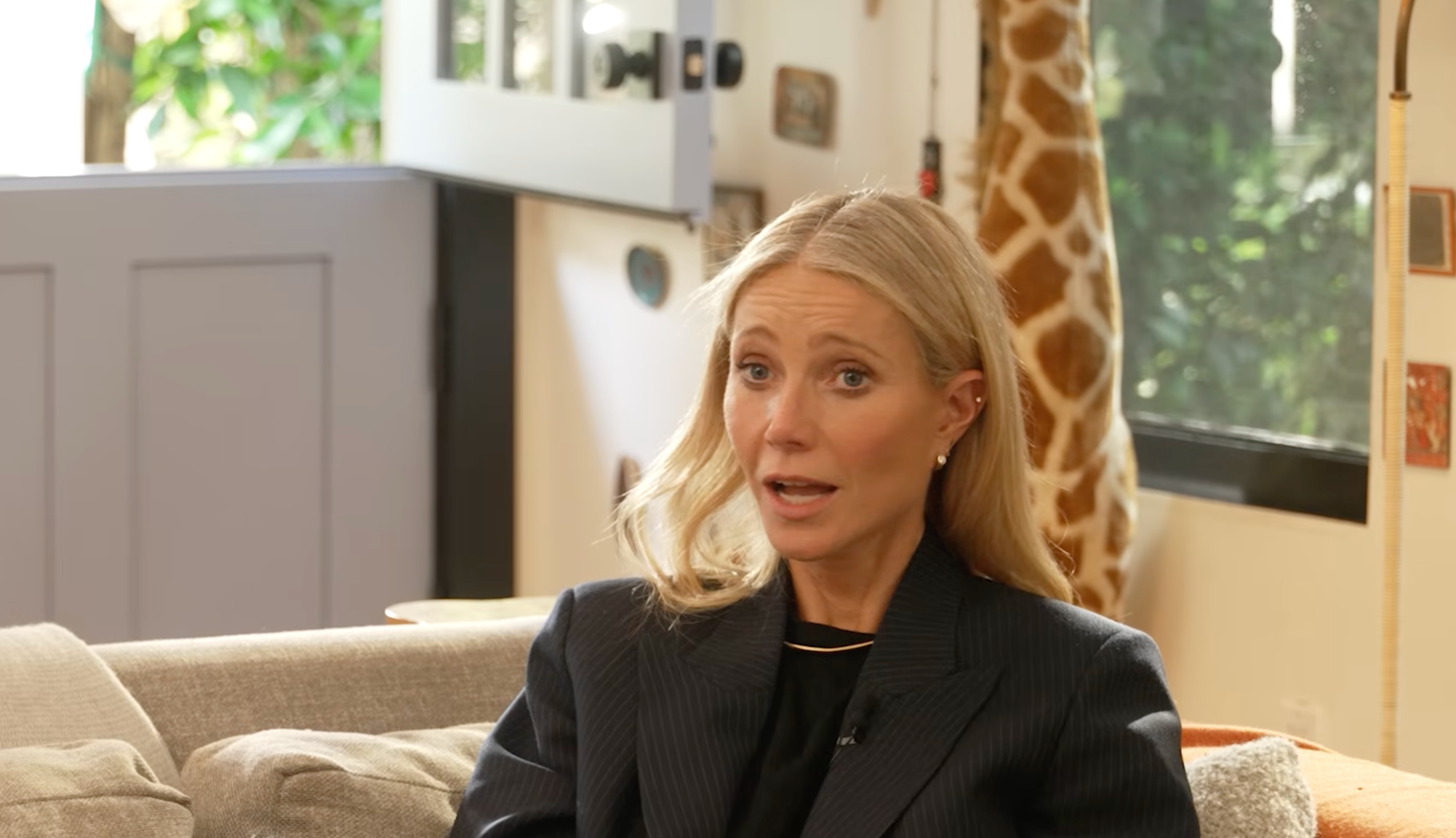 Gwyneth Paltrow sitting on a couch in a cozy room, wearing a dark blazer, with a giraffe decoration in the background.
