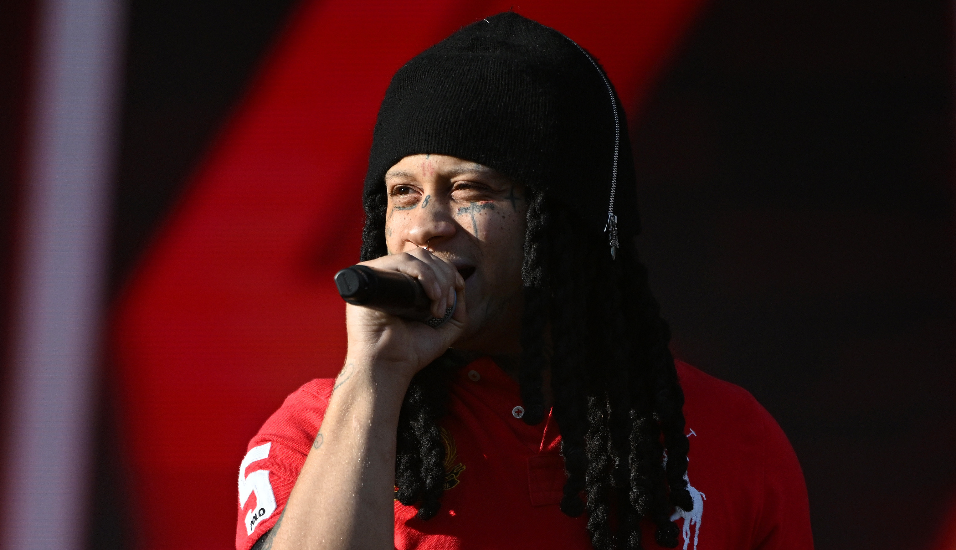 Trippie Redd with long hair and a beanie sings into a microphone, wearing a red "Milan" polo shirt on stage.