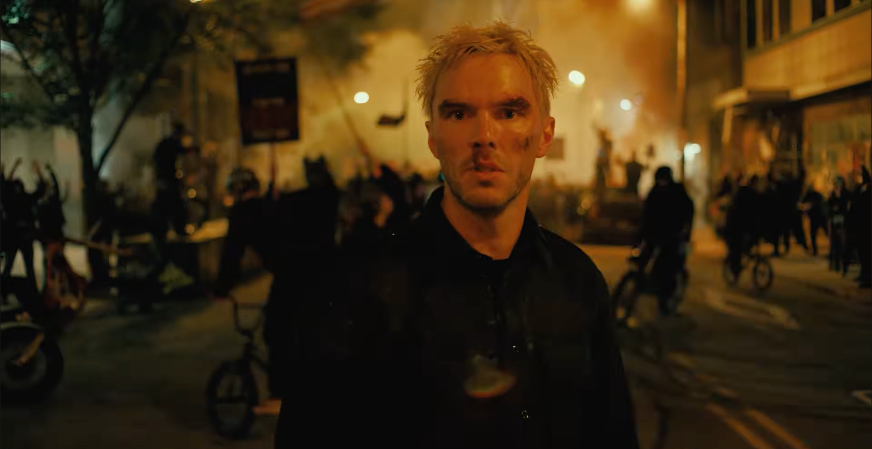 A man with blond hair stands in a chaotic street scene at night, with people and smoke in the background.